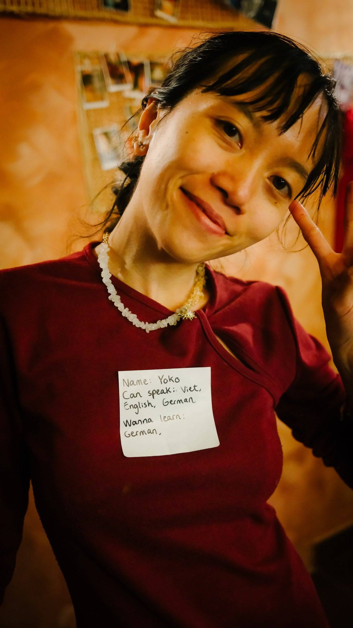 A woman smiles warmly in a cozy indoor setting with a name tag indicating she speaks Vietnamese and English, and is learning German at an event in Đà Lạt.