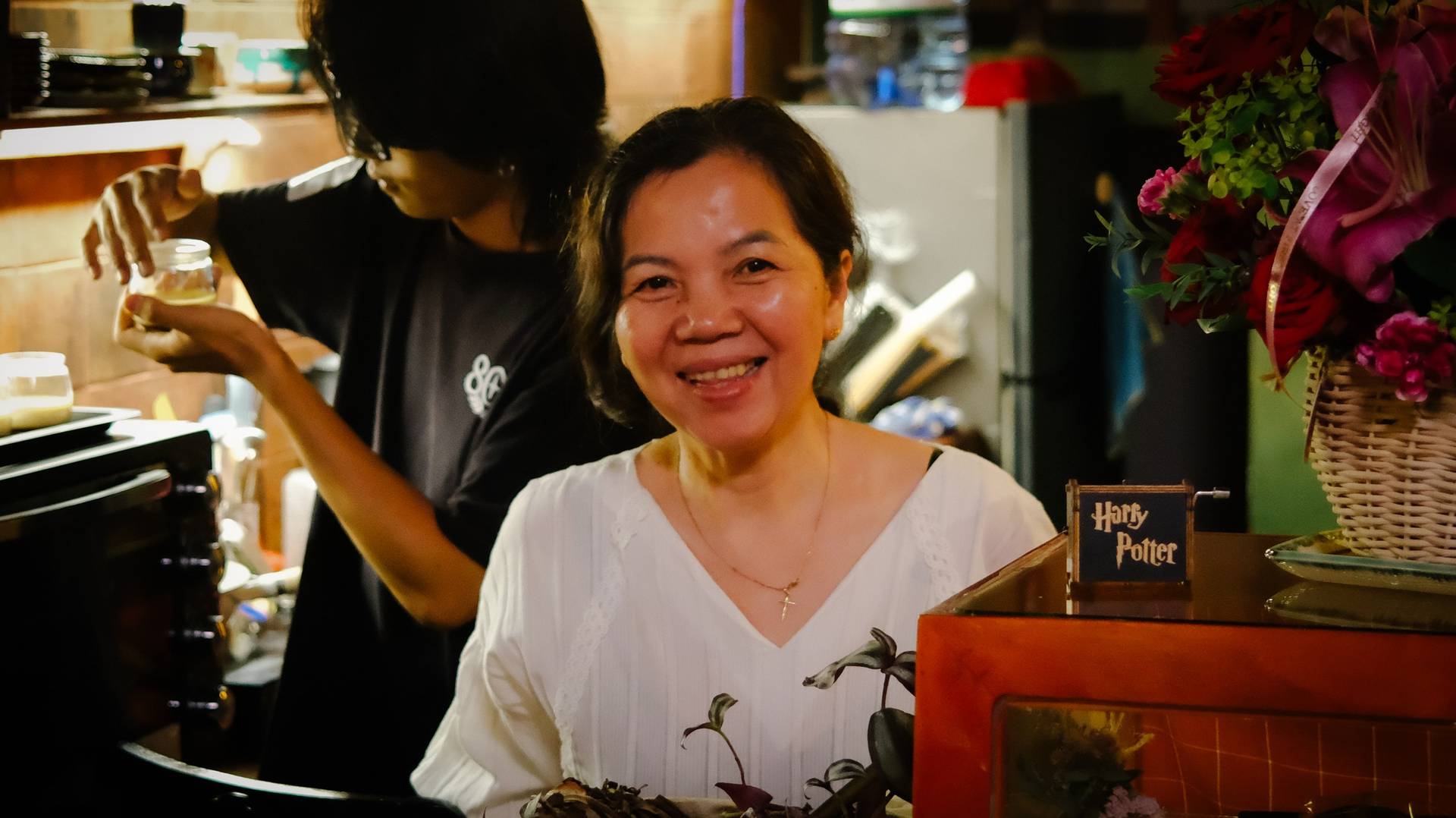 A cheerful woman smiles warmly in a cozy café setting in Đà Lạt, Vietnam, surrounded by floral decorations and a Harry Potter-themed display.