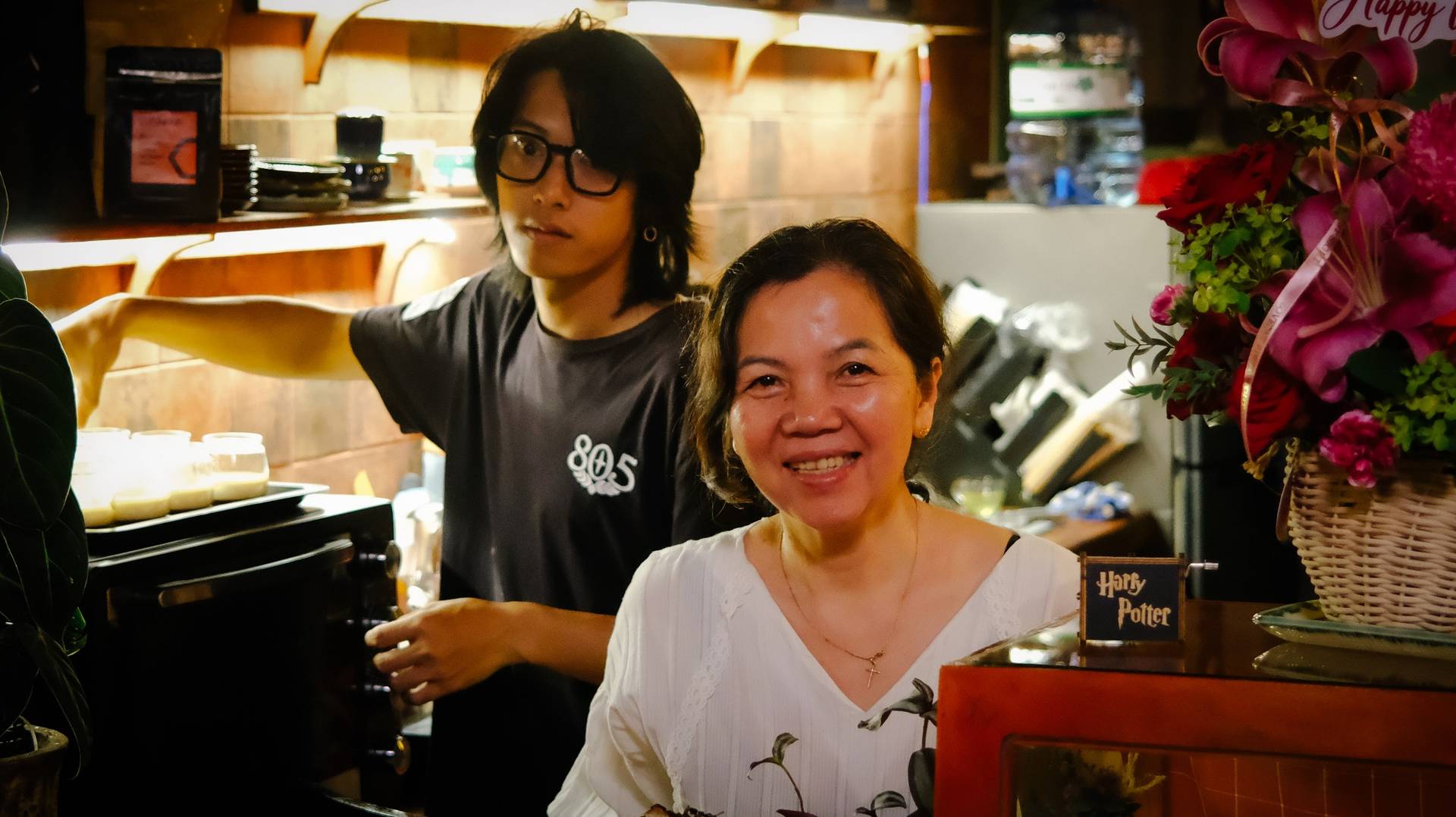 A cheerful moment in a cozy café setting in Đà Lạt, Vietnam, where two individuals share a warm smile amidst a backdrop of floral decorations and coffee-making equipment.