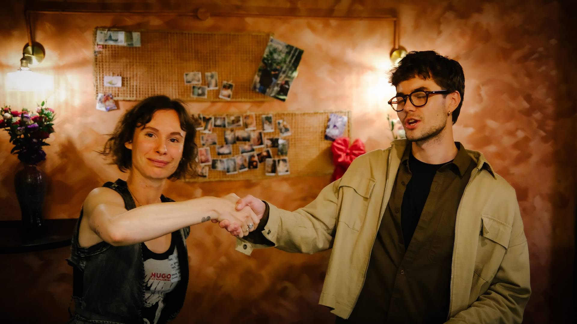 Two individuals share a warm handshake in a cozy, warmly lit room adorned with personal photos and flowers, suggesting a friendly or celebratory gathering.