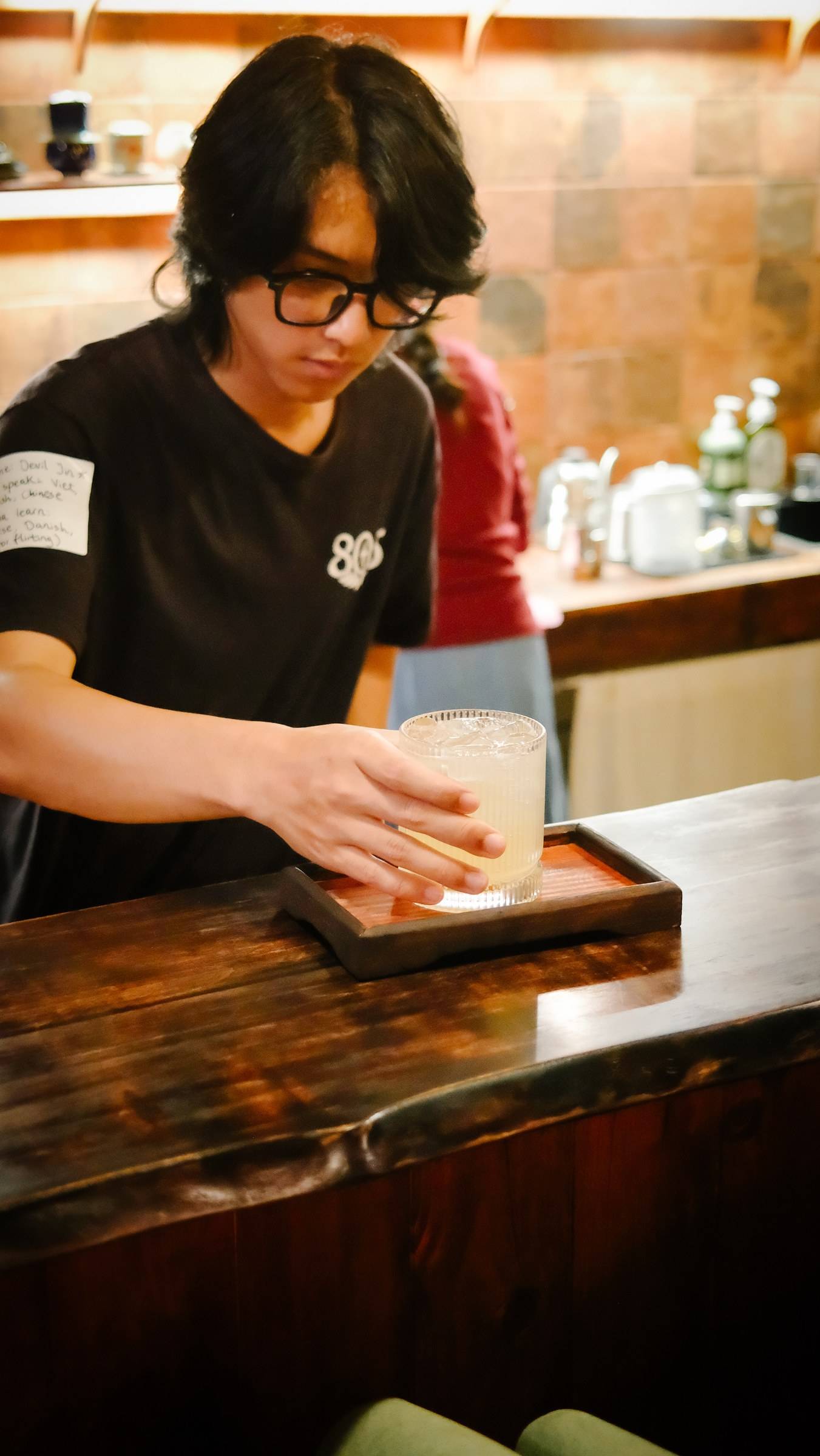 A bartender in Đà Lạt carefully serves a refreshing drink with ice cubes to a customer seated at a wooden bar counter.