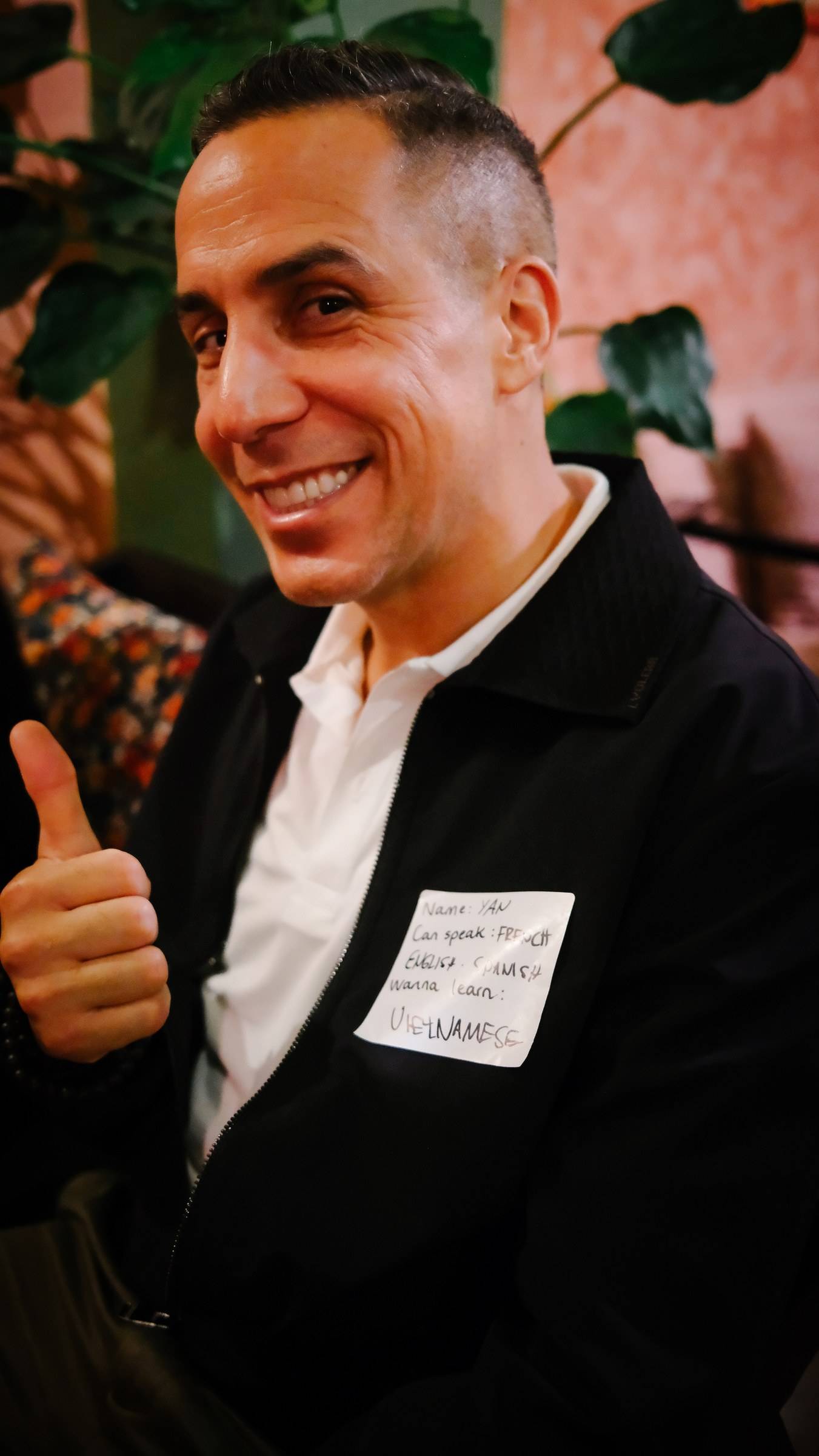 A person smiling warmly with a thumbs-up gesture in front of a vibrant floral backdrop, indicating a friendly and welcoming atmosphere at an event in Đà Lạt.