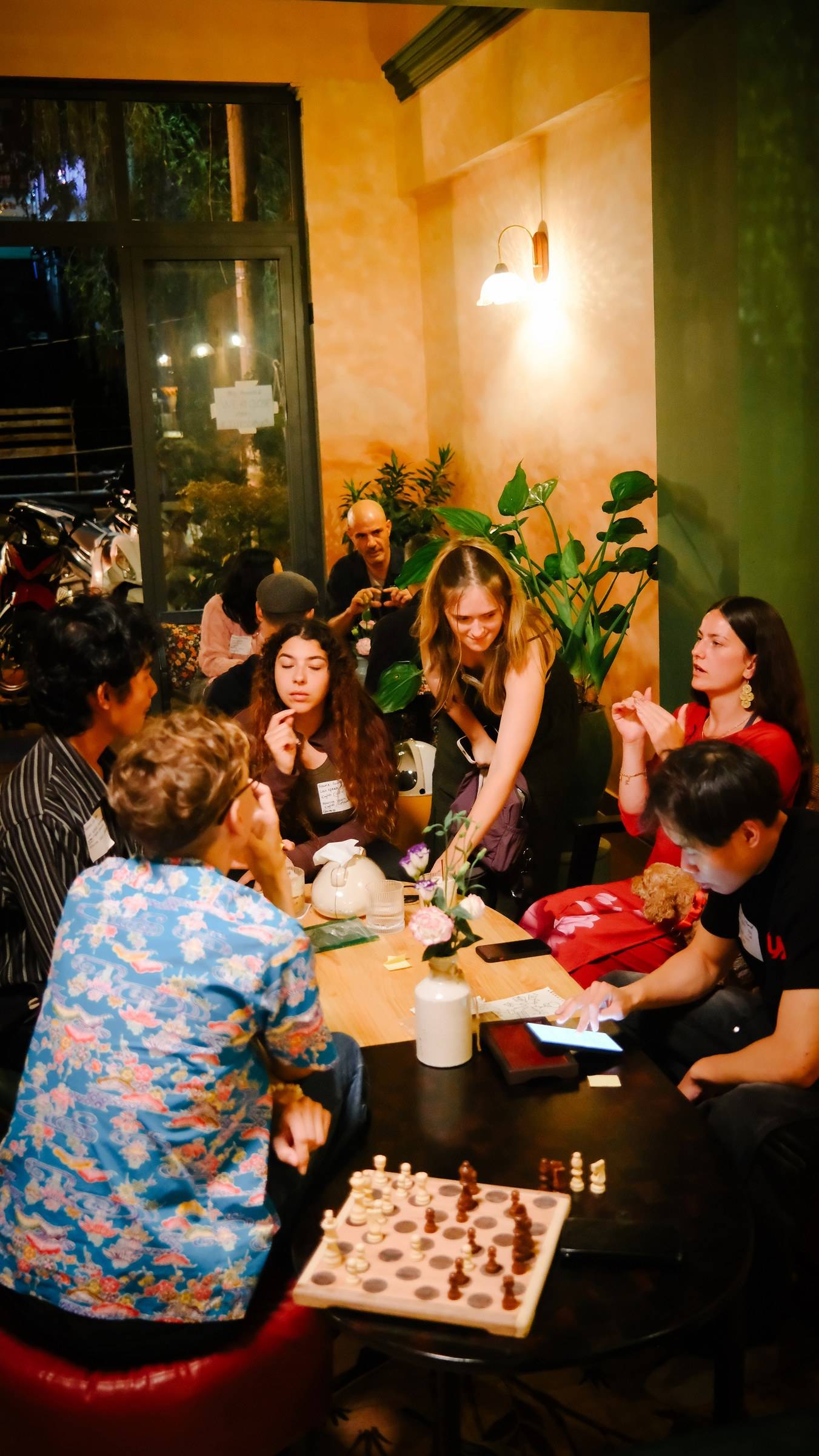 A group of friends gathered in a cozy café in Đà Lạt, Vietnam, engaging in lively conversation and playing chess at a wooden table adorned with flowers.