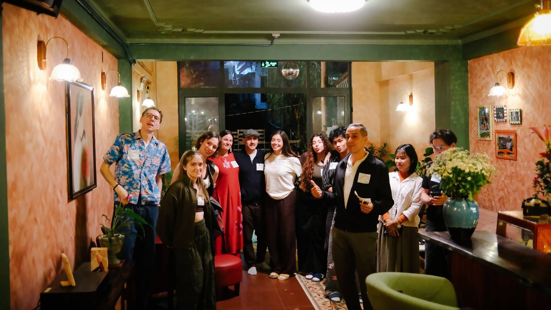 A group of friends gather in a cozy, warmly lit room with vibrant decor and floral arrangements, enjoying each other's company at an event in Đà Lạt.