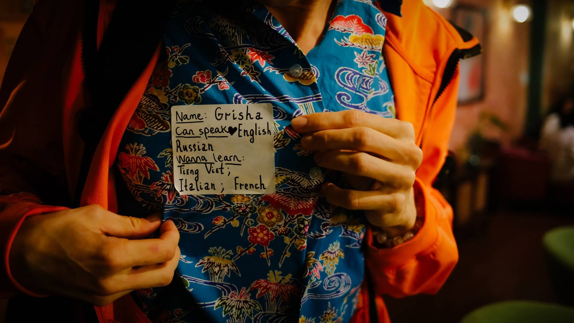 A person in a vibrant floral shirt holds up a note indicating they can speak English and Russian while wanting to learn Vietnamese, Italian, and French at an event in Đà Lạt.