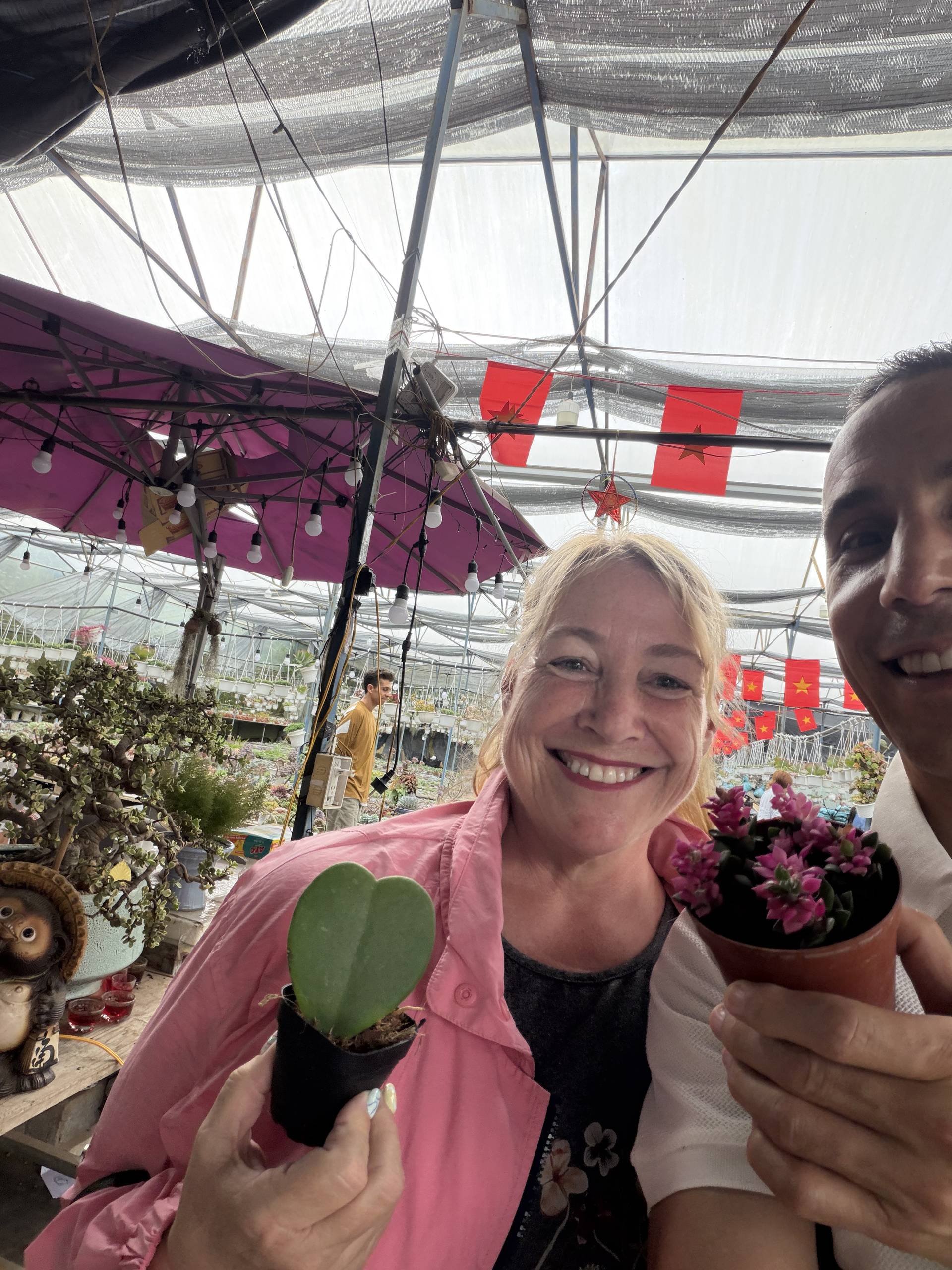 Two individuals are enjoying a sunny day in Đà Lạt's greenhouse market, holding small potted plants and smiling amidst vibrant greenery and Vietnamese flags.
