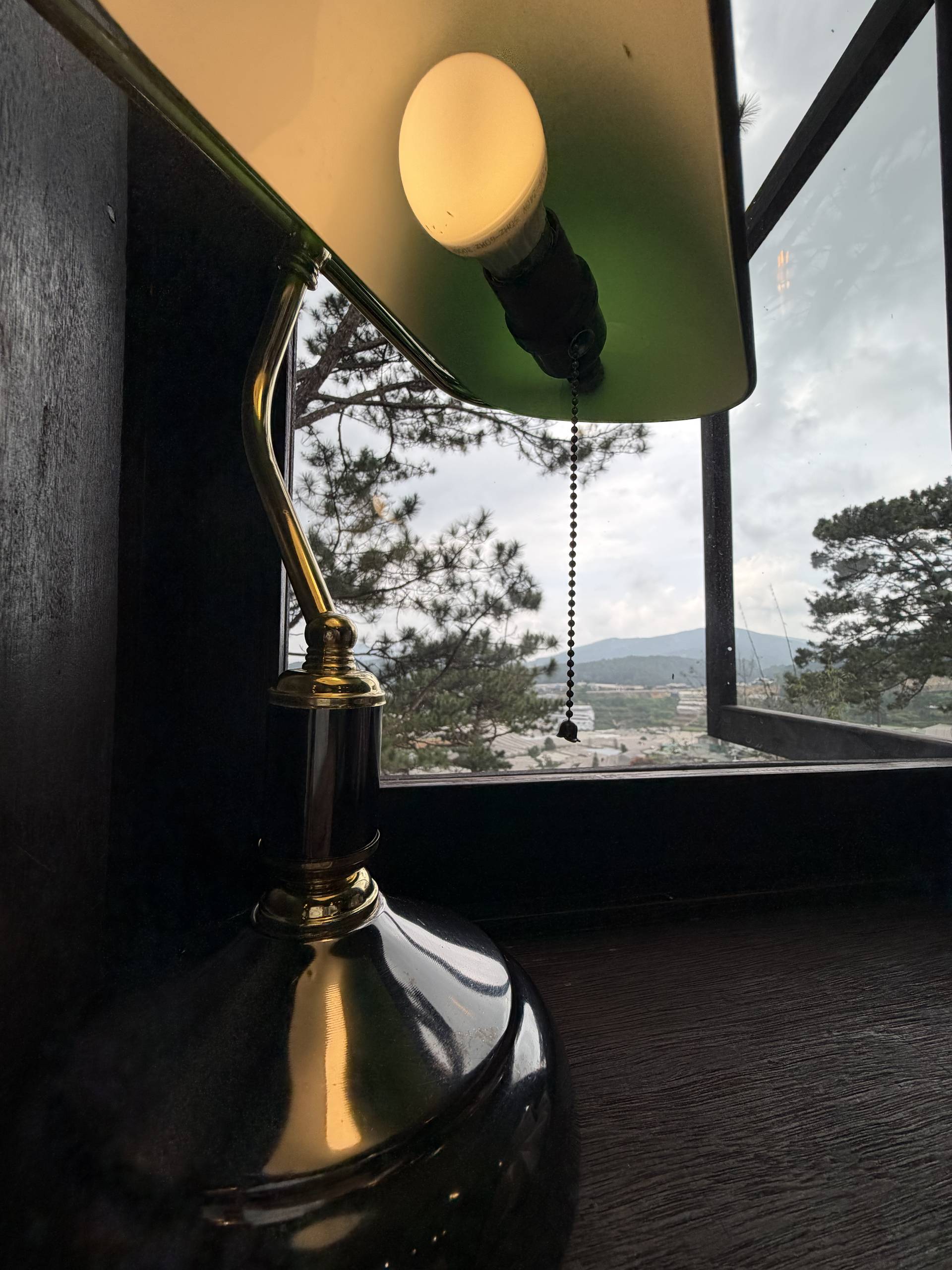 A vintage-style desk lamp with a green shade sits by a window in Đà Lạt, Vietnam, offering a serene view of pine trees and distant hills under an overcast sky.