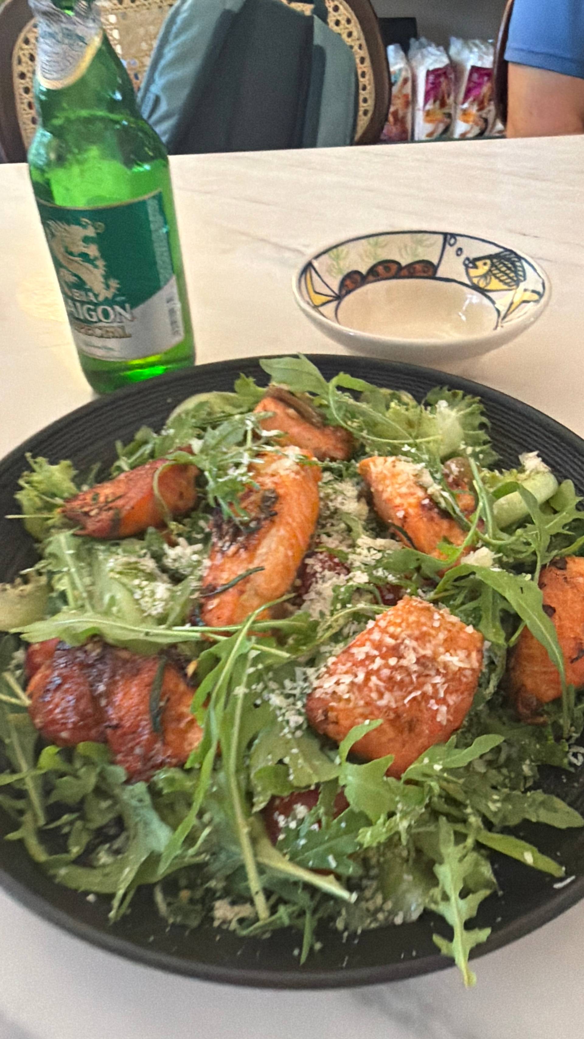 A vibrant salad with grilled salmon, arugula, and Parmesan cheese is served in a cozy café setting in Đà Lạt, Vietnam, accompanied by a bottle of beer.