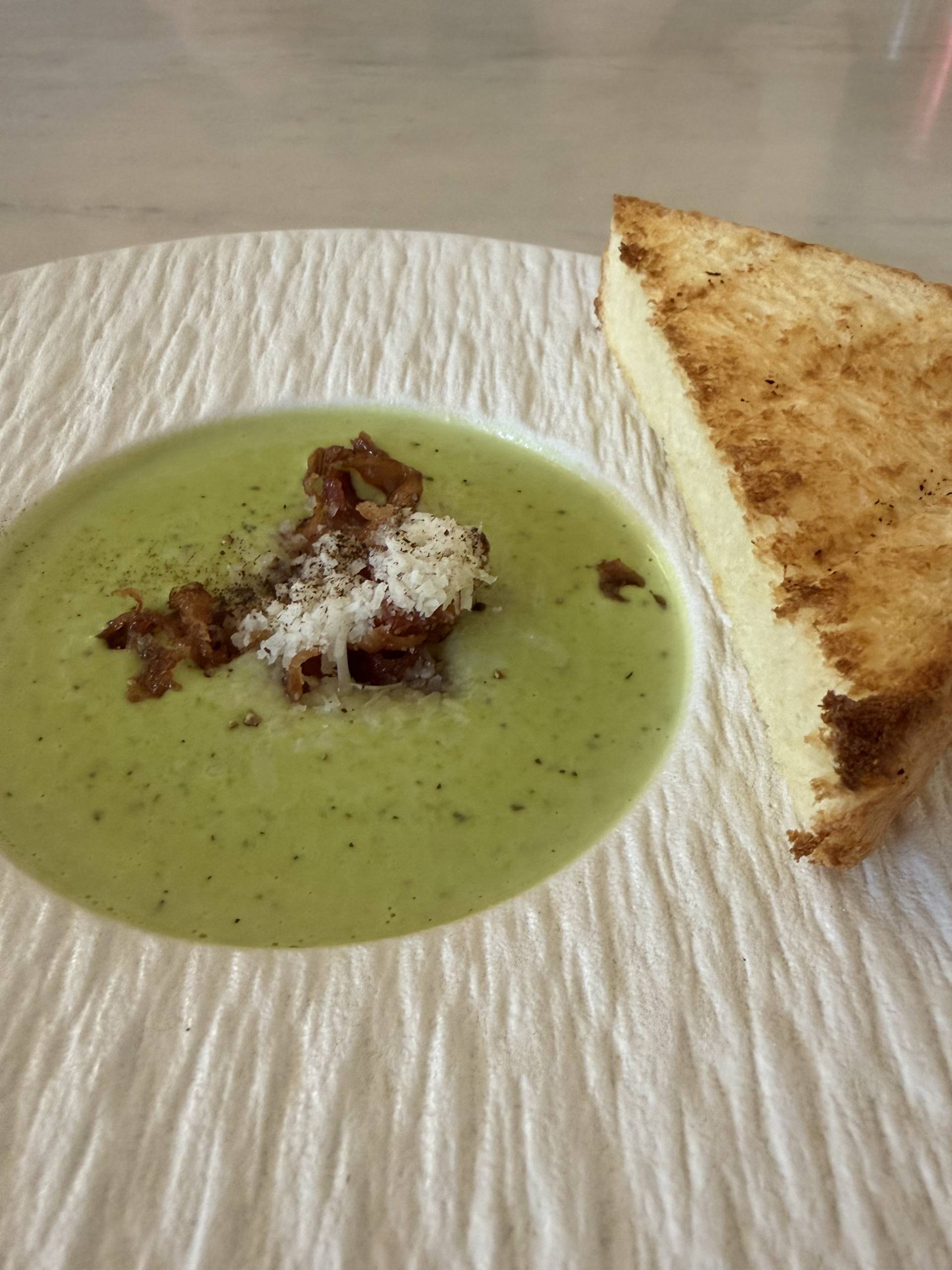 A creamy green soup garnished with crispy bacon bits and grated cheese sits elegantly in a white bowl beside a slice of toasted bread, suggesting a cozy and comforting meal setting.