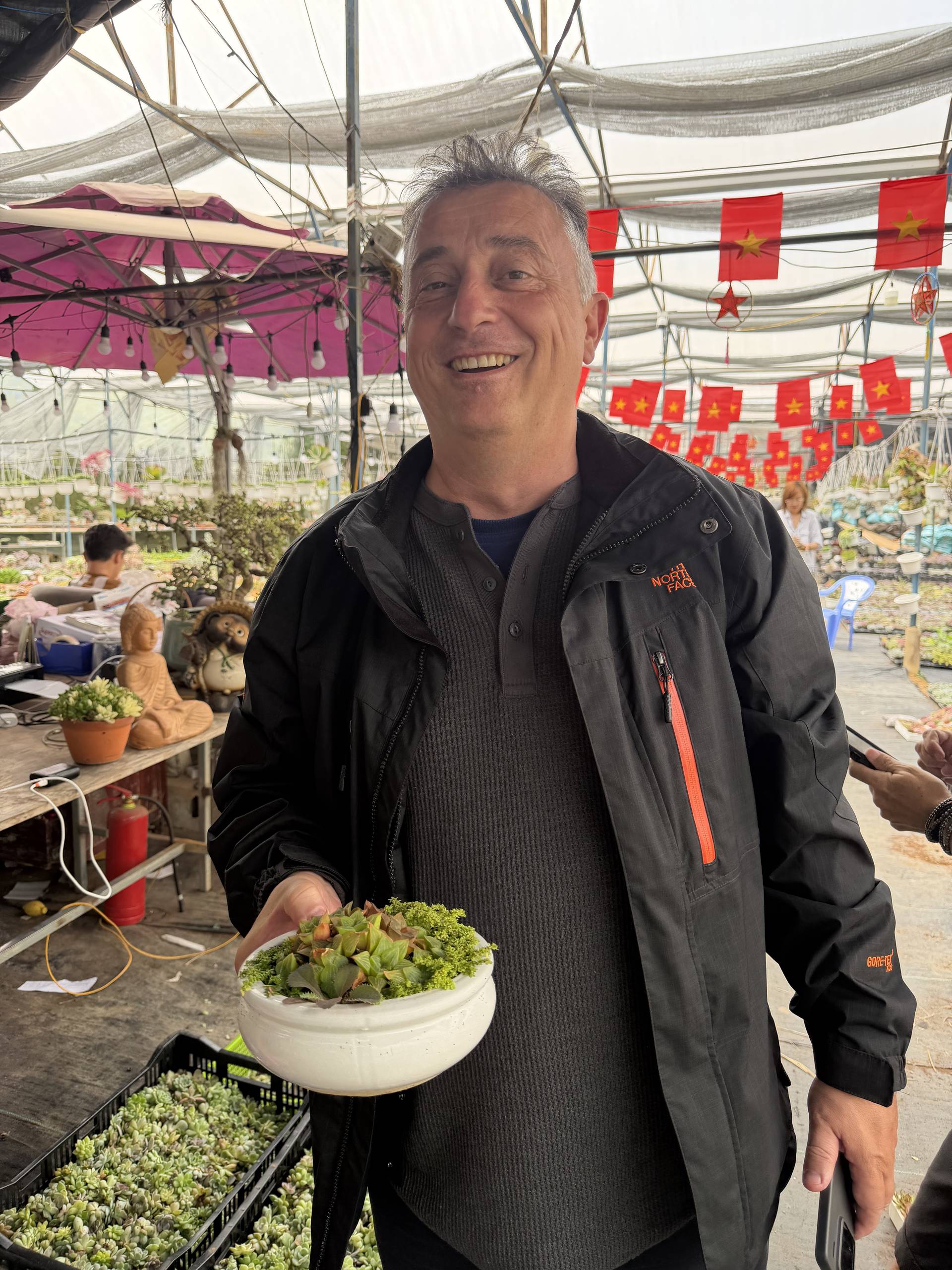 A man smiles while holding a small succulent arrangement in a market setting adorned with Vietnamese flags and greenery, reflecting a vibrant and cheerful atmosphere.