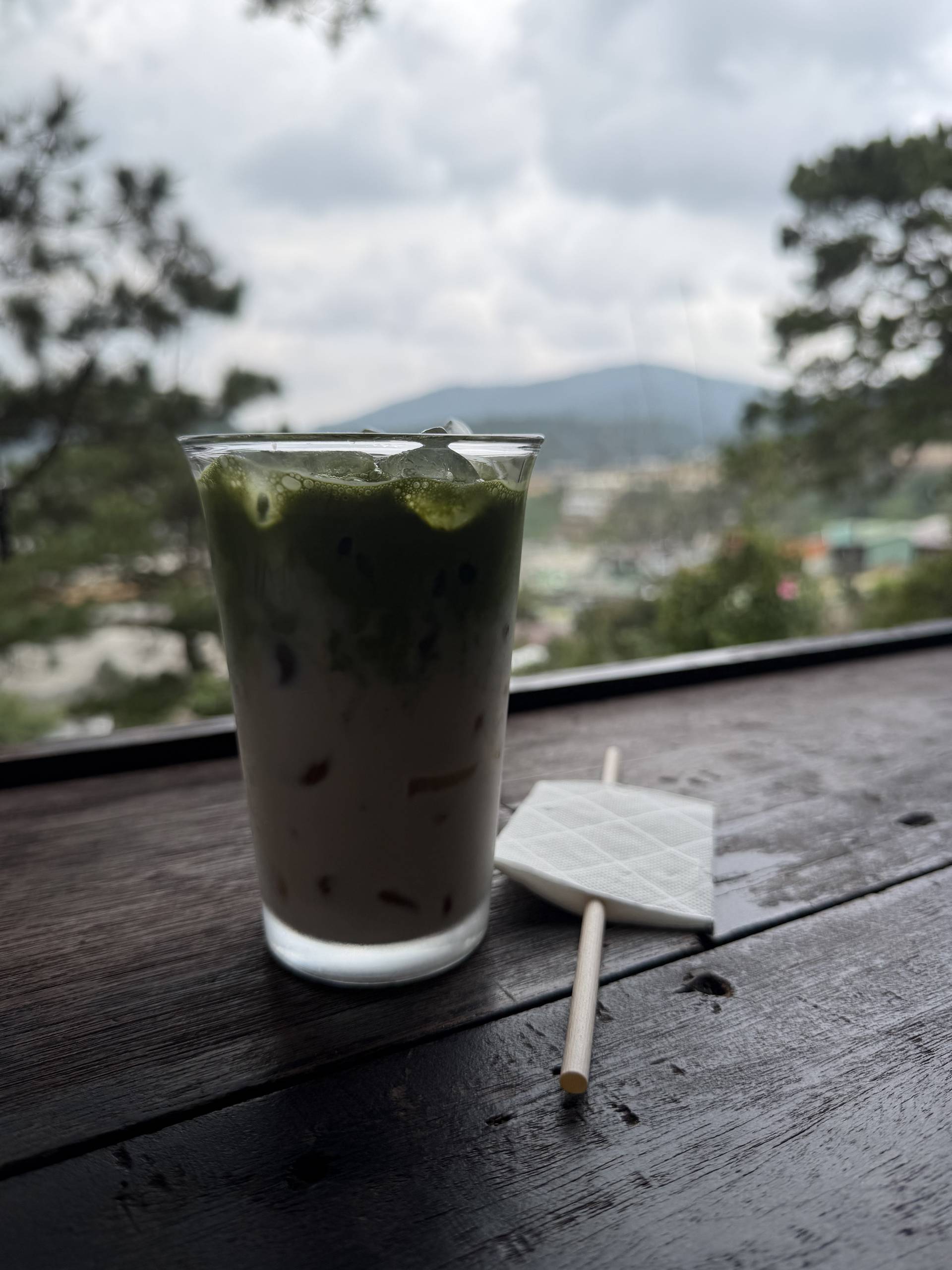 A refreshing iced matcha latte with ice cubes sits on a wooden table against a scenic backdrop of lush greenery and distant mountains in Đà Lạt, Vietnam.