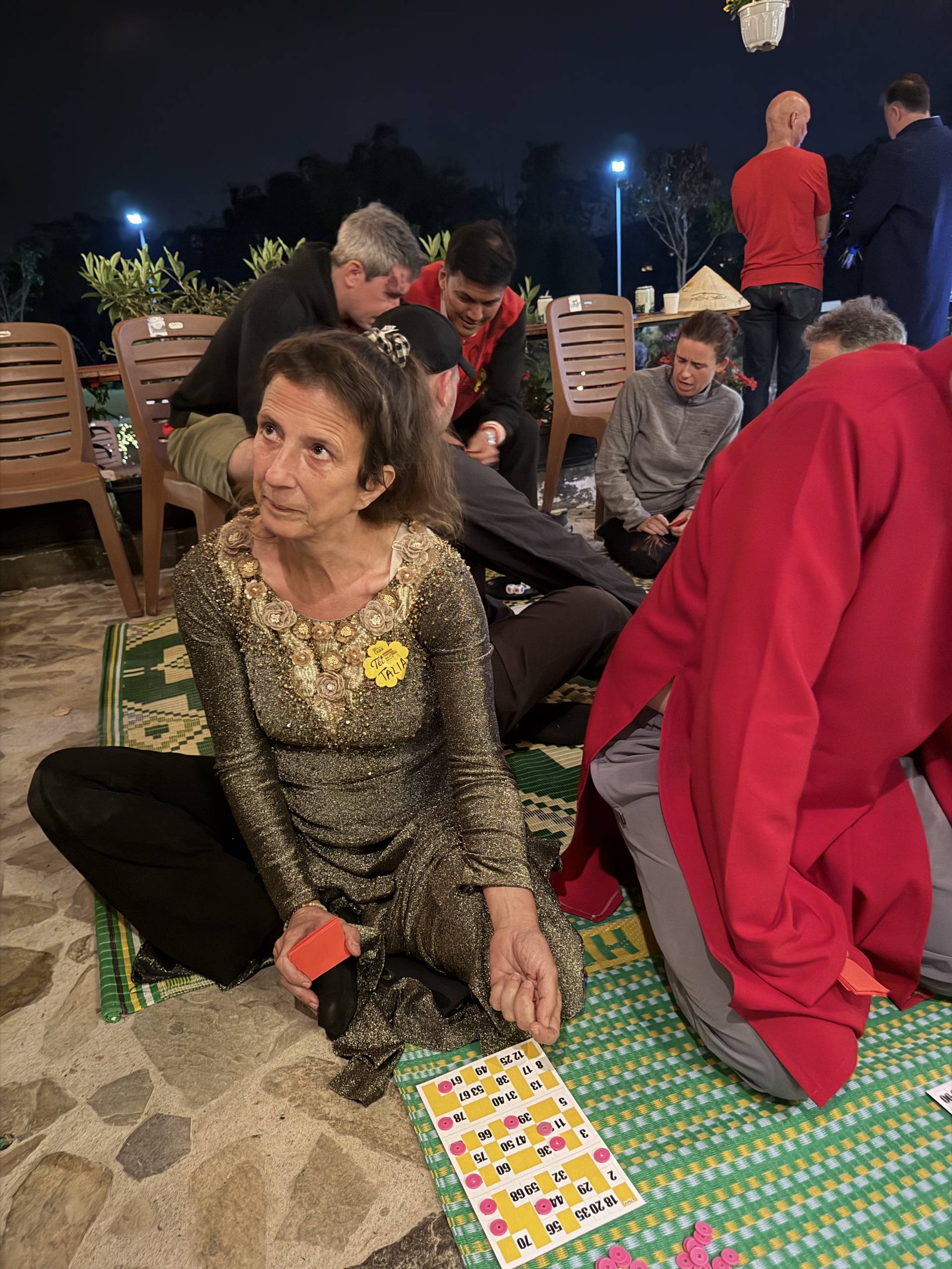 Evening social gathering in Đà Lạt under string lights and lanterns. Groups enjoy bingo and games on patterned mats surrounded by potted flowers and warm nighttime ambiance.