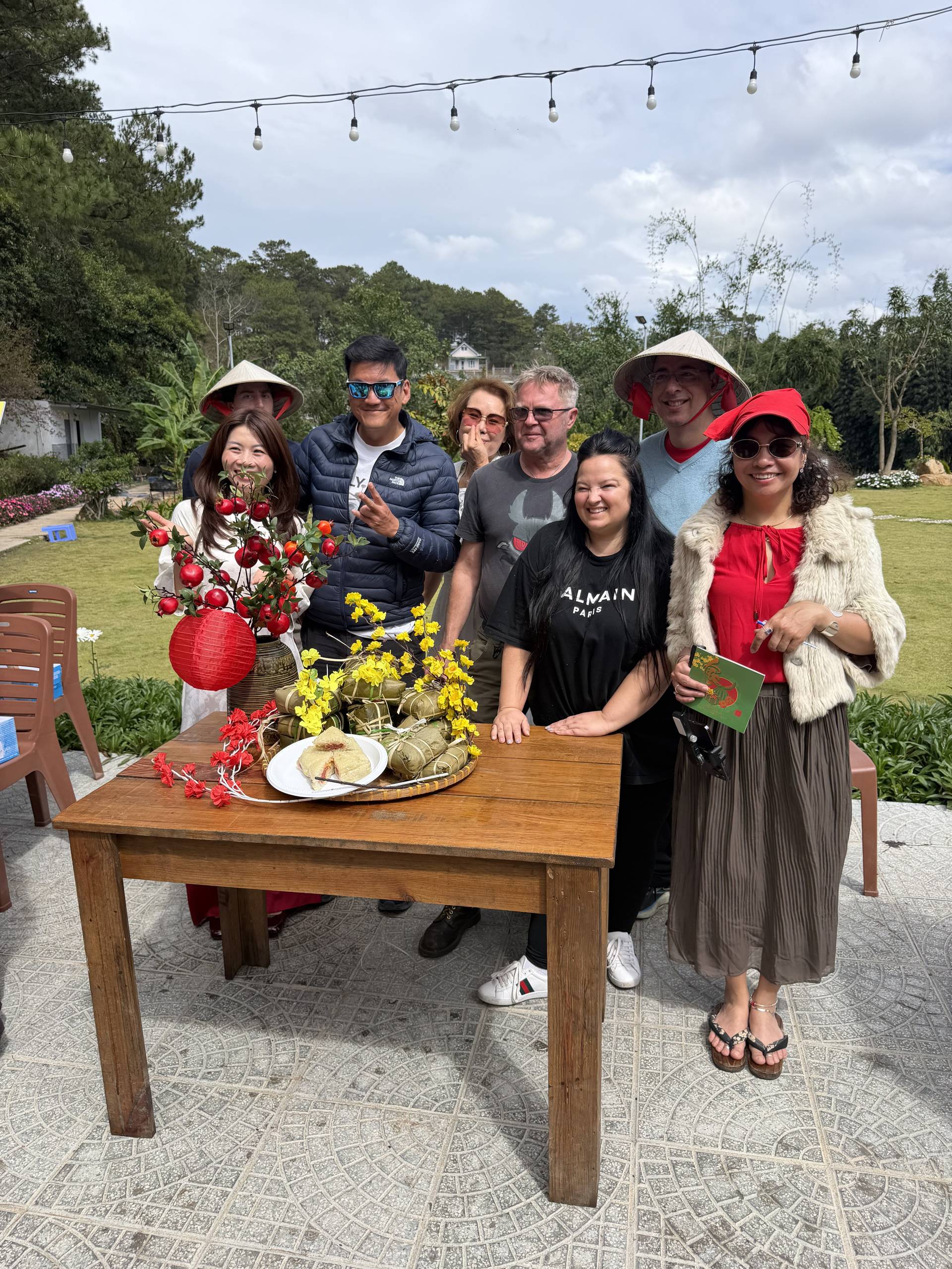 Festive outdoor gathering celebrating Vietnamese culture in Đà Lạt. A group shares traditional food and flowers around a rustic wooden table with string lights, conical hats, and pine forest views.