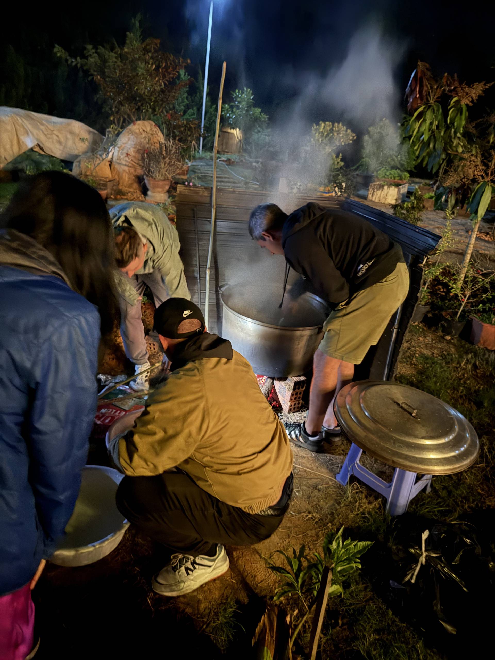 Nighttime communal cooking event in Đà Lạt featuring a large pot of food preparation outdoors, surrounded by participants in a pine forest garden setting, capturing traditional Vietnamese hospitality and outdoor gathering culture.