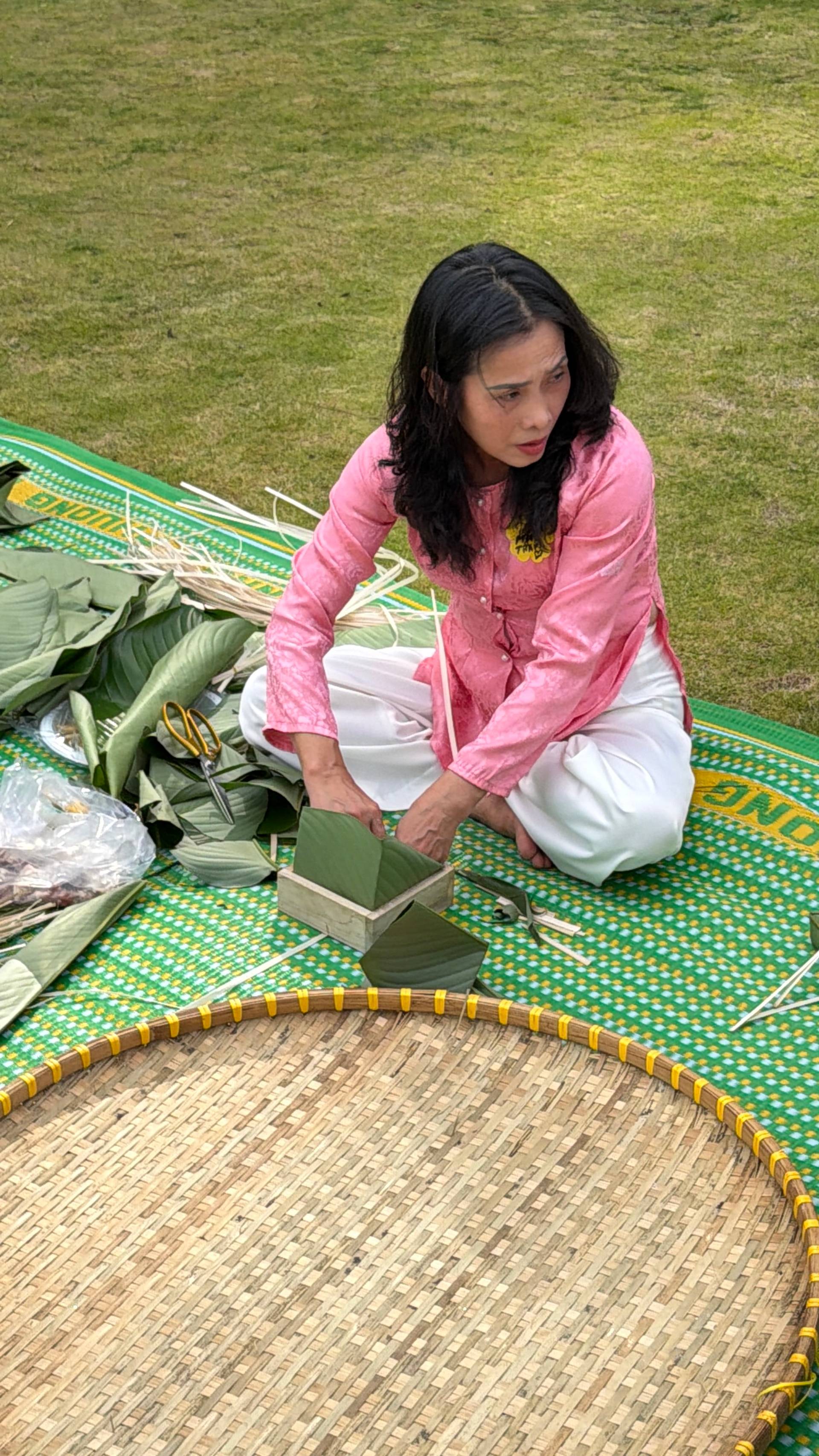 Traditional Vietnamese craft workshop at a Đà Lạt event, showing hands-on participation in bamboo or palm leaf weaving on a decorated mat, set against a lush green lawn typical of the city's garden spaces.