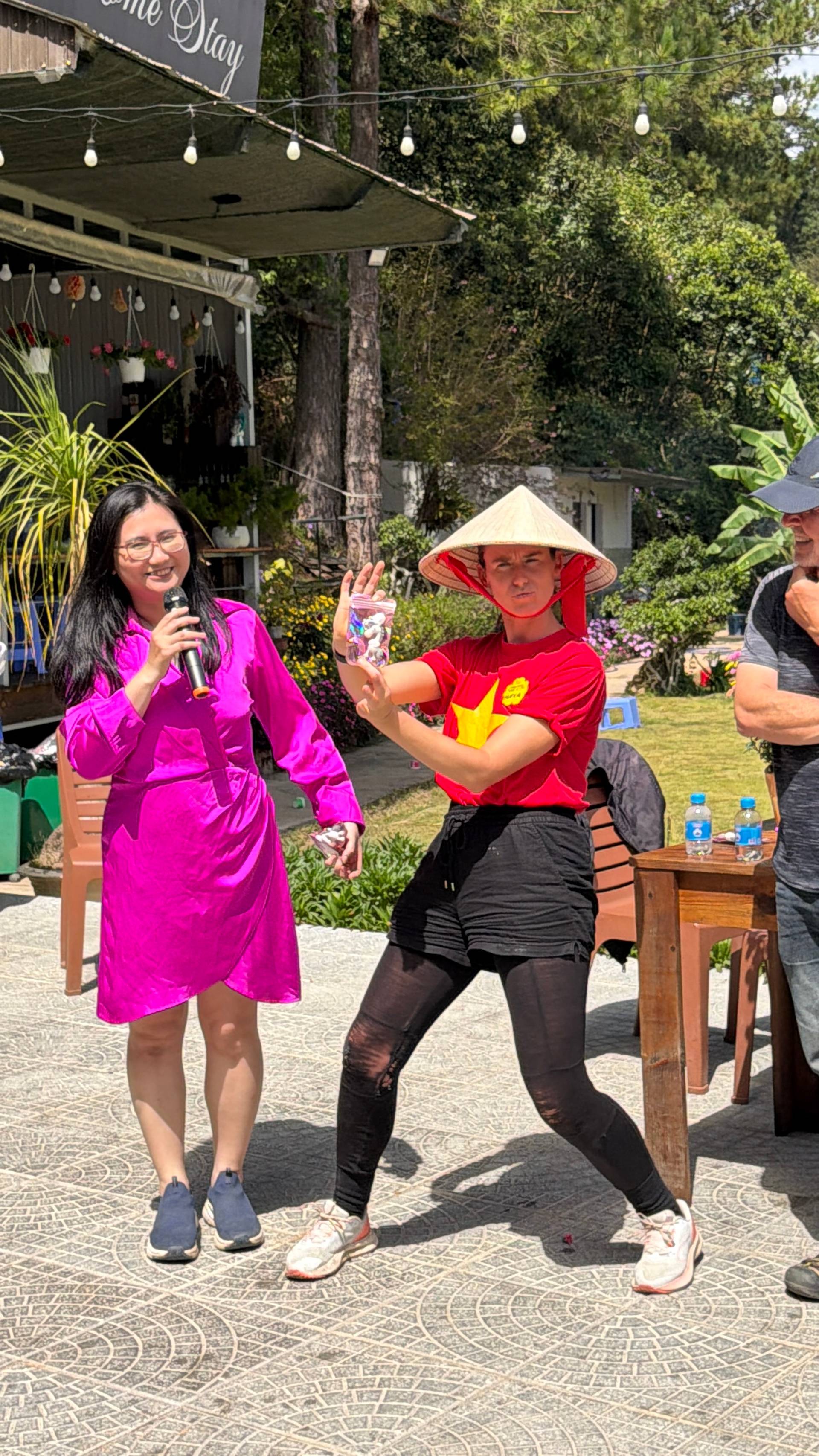 Performer in vibrant magenta áo dài holds microphone during entertainment segment at Đà Lạt festival, surrounded by traditional nón lá hats, colorful flowers, and wooden event infrastructure.