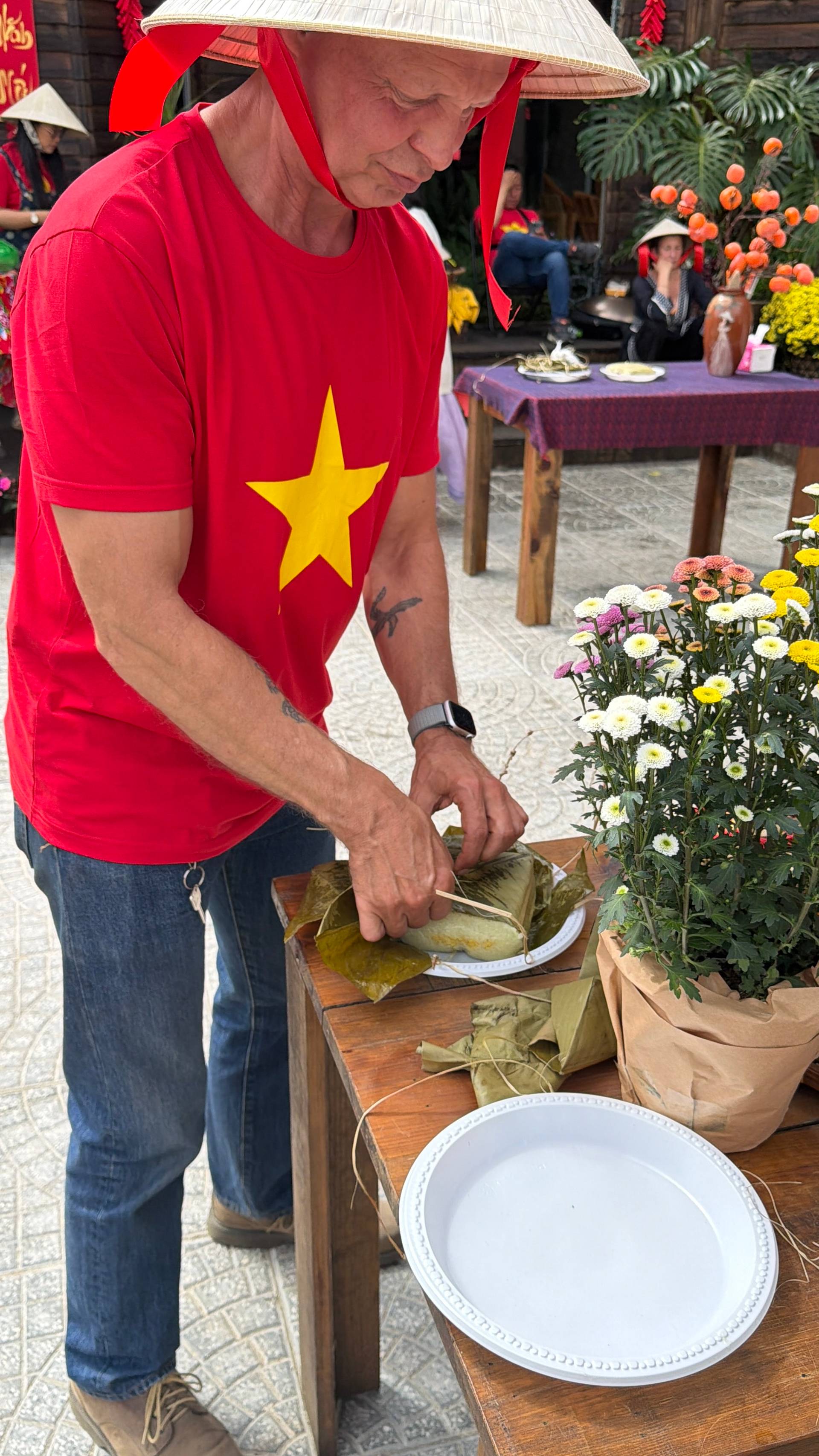 Festival attendee in Vietnamese flag shirt and traditional nón lá hat serves wrapped bánh chưng among vibrant carnations and chrysanthemums at Đà Lạt cultural event.