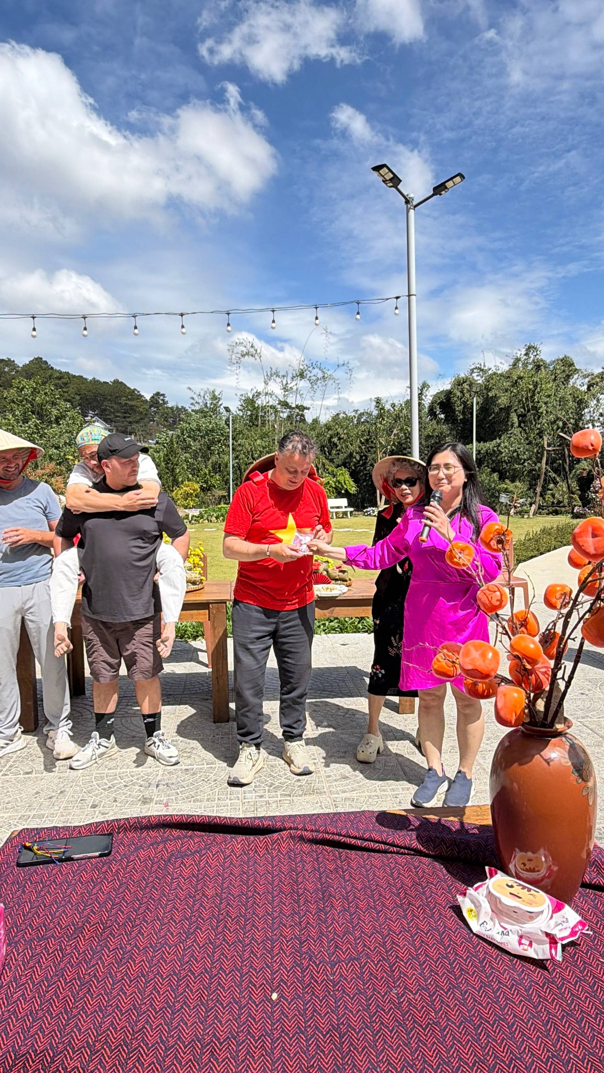 Outdoor Đà Lạt festival gathering under clear blue sky with string lights, decorative gourds, and vibrant flowers surrounding traditional tables in pine-forested highland venue.