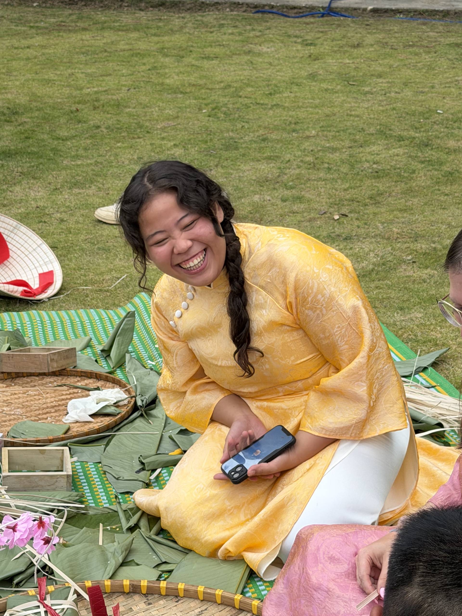 Joyful attendee in golden áo dài sits on woven mat during Đà Lạt cultural event, holding traditional crafts and smiling amid festive highland garden setting.