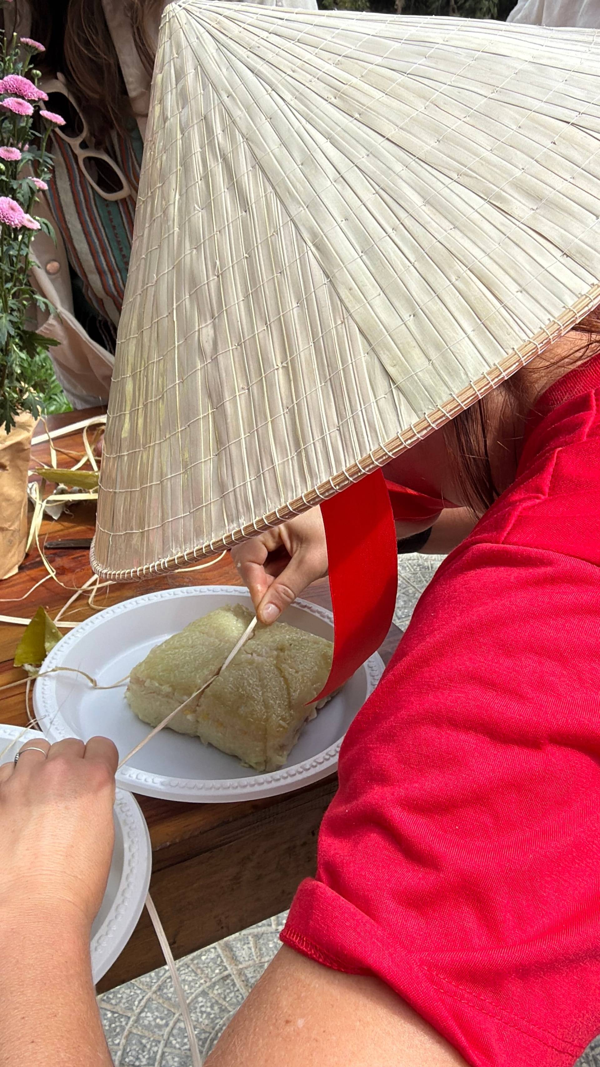 Traditional Vietnamese conical hat held over wrapped bánh chưng mooncakes at Đà Lạt festival, surrounded by pink flowers and festive cultural decor.