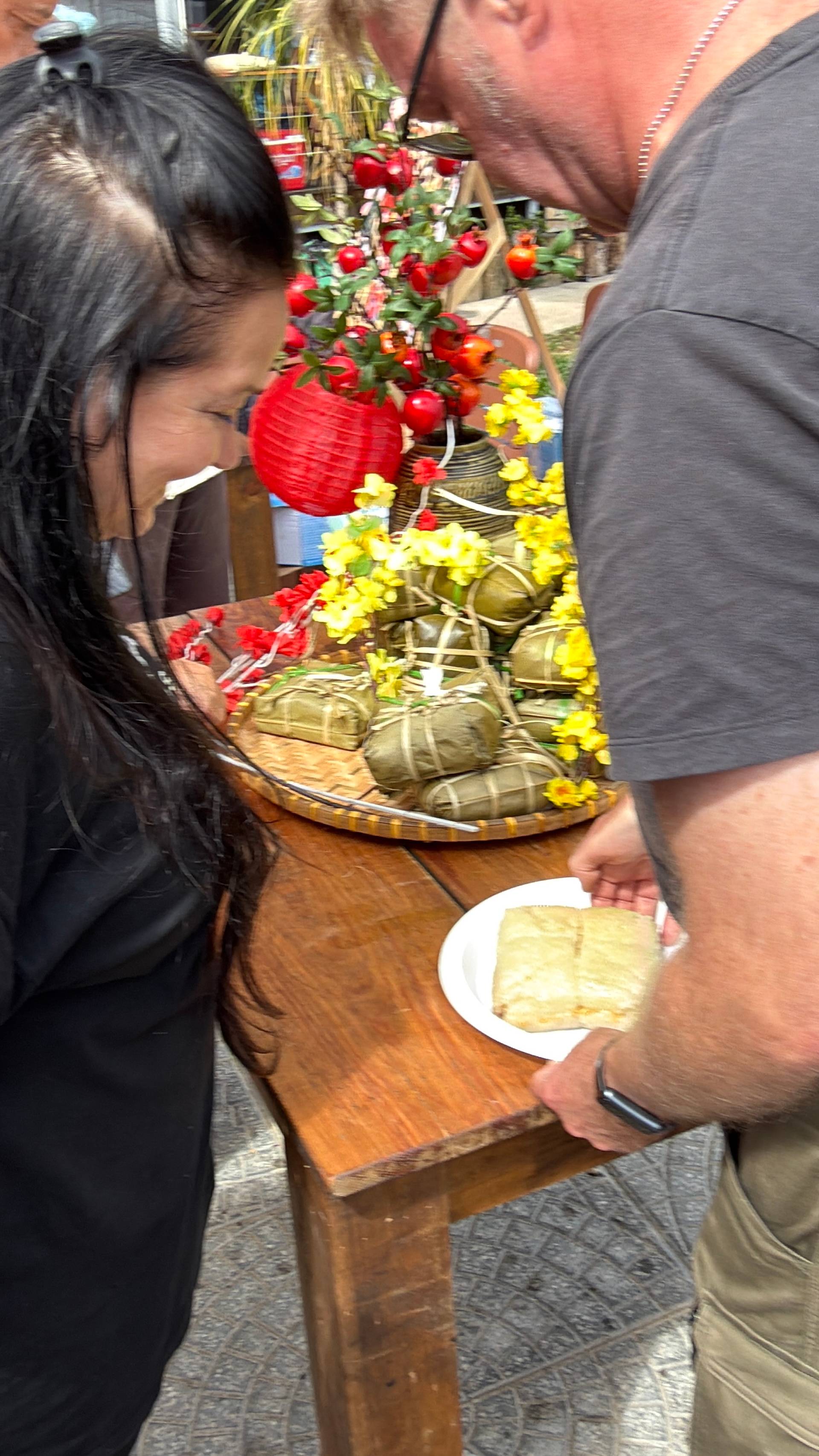 Close-up of traditional Vietnamese food tasting at Đà Lạt event, featuring bánh chưng on woven platter surrounded by red lanterns and vibrant festival flowers.