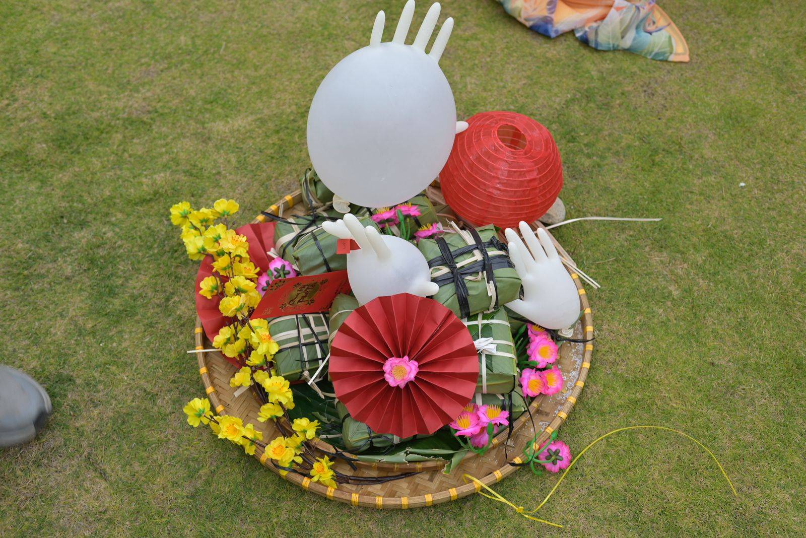 Overhead aerial view of decorative floral arrangement on Đà Lạt lawn featuring traditional Vietnamese craft elements. Circular design with white hand shapes, red paper lanterns, and vibrant yellow and pink flowers.