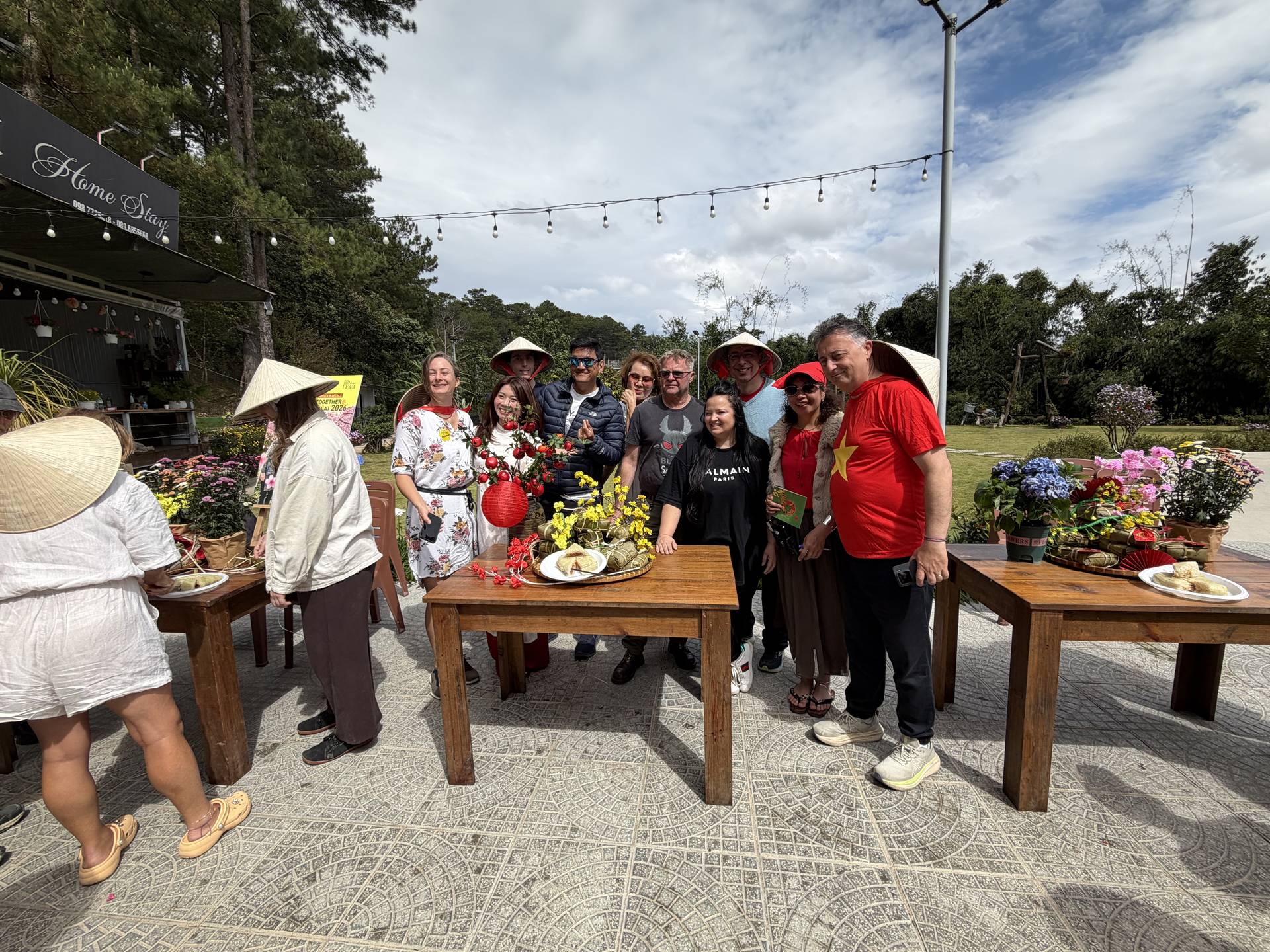 Group celebration at Dalat Stay garden venue with large gathering in traditional and formal attire. Daytime event featuring red-themed decorations, pine forest backdrop, and decorative string lights creating festive atmosphere.