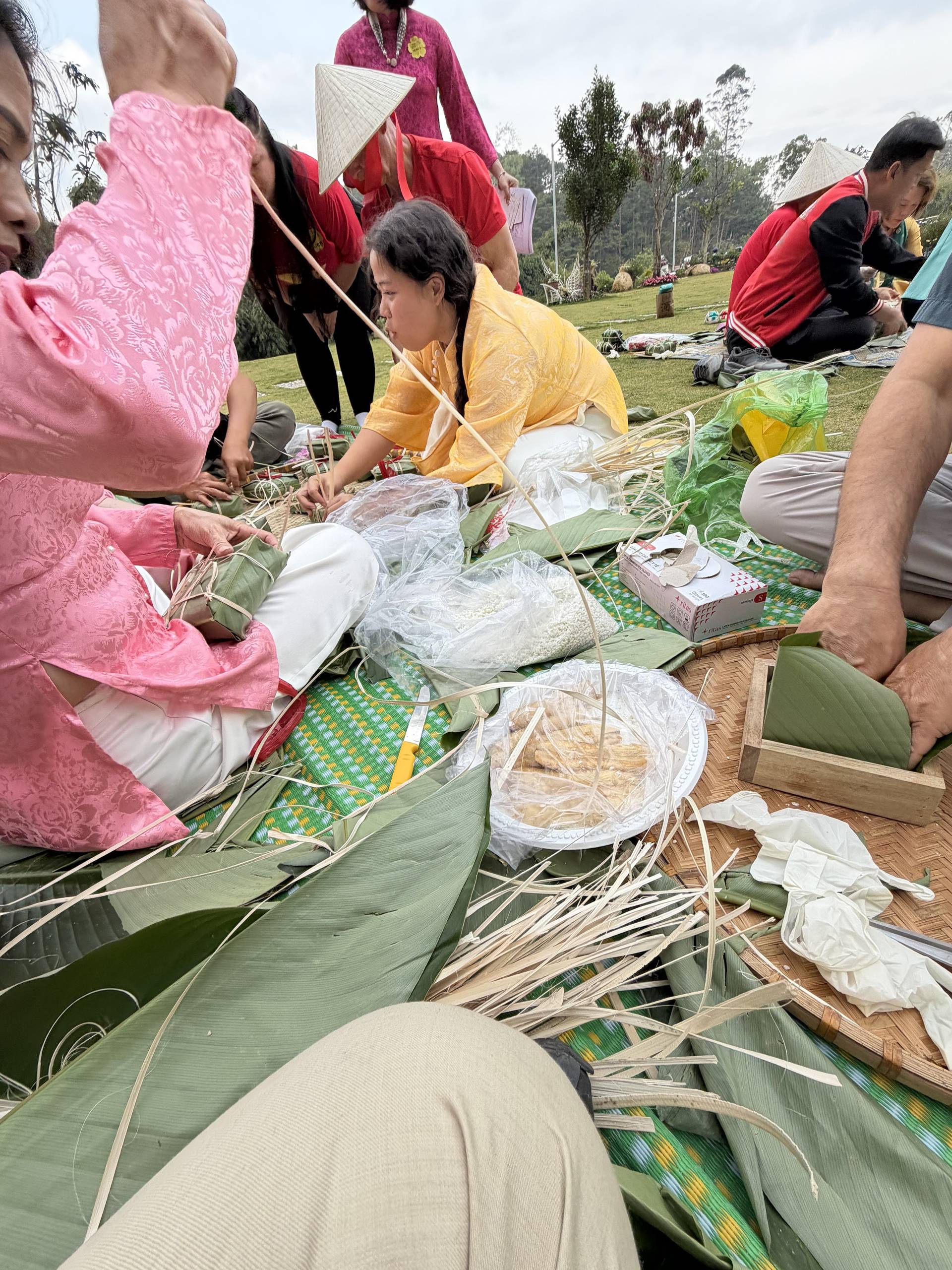 Group workshop at a Đà Lạt event featuring traditional conical hat weaving, with participants in colorful áo dài actively crafting on a hillside garden surrounded by pine trees.