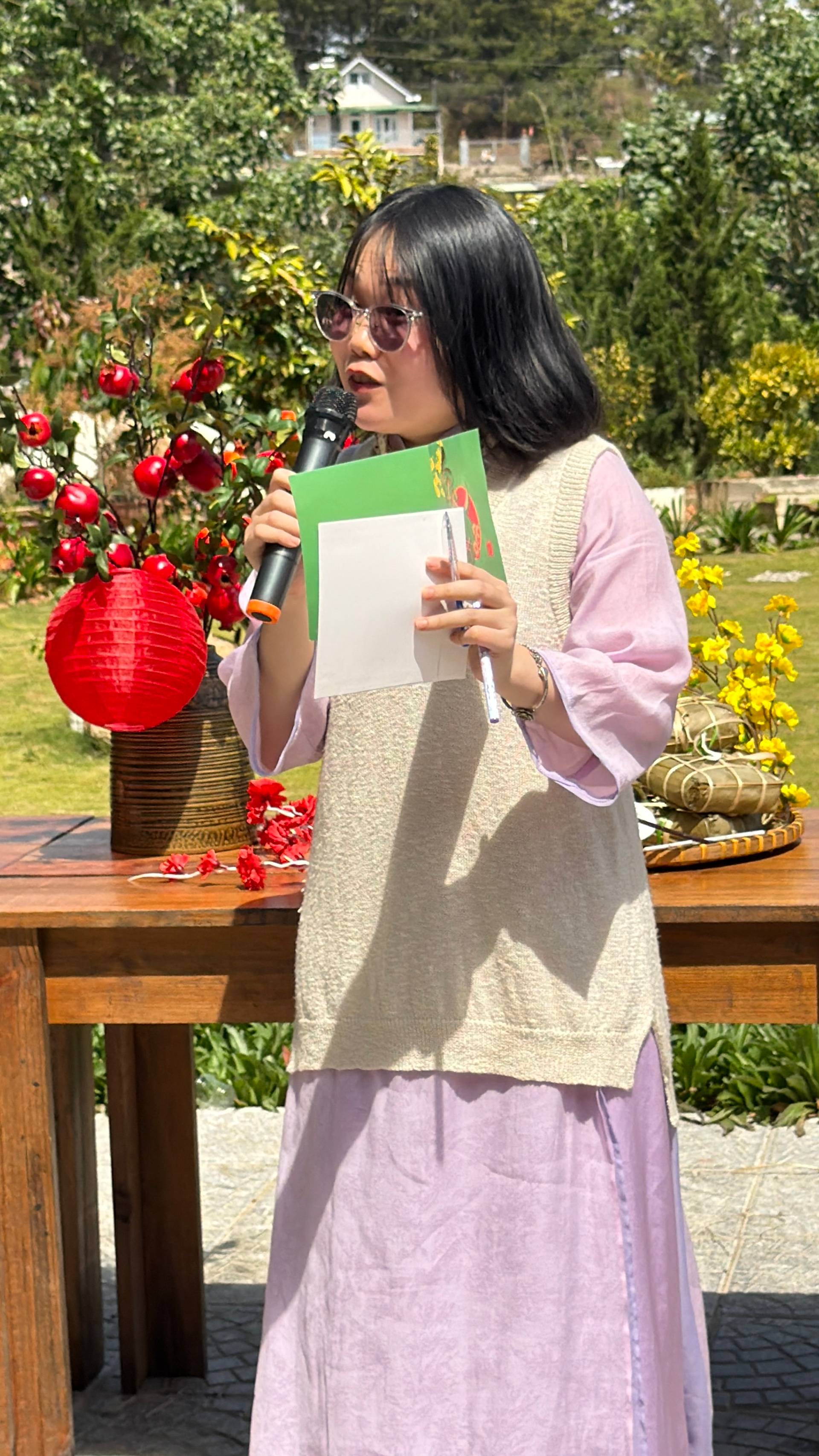 Presenter speaking at outdoor Đà Lạt event with microphone and program in hand. Garden celebration features red rose arrangements and lush pine forest setting typical of Đà Lạt highlands.