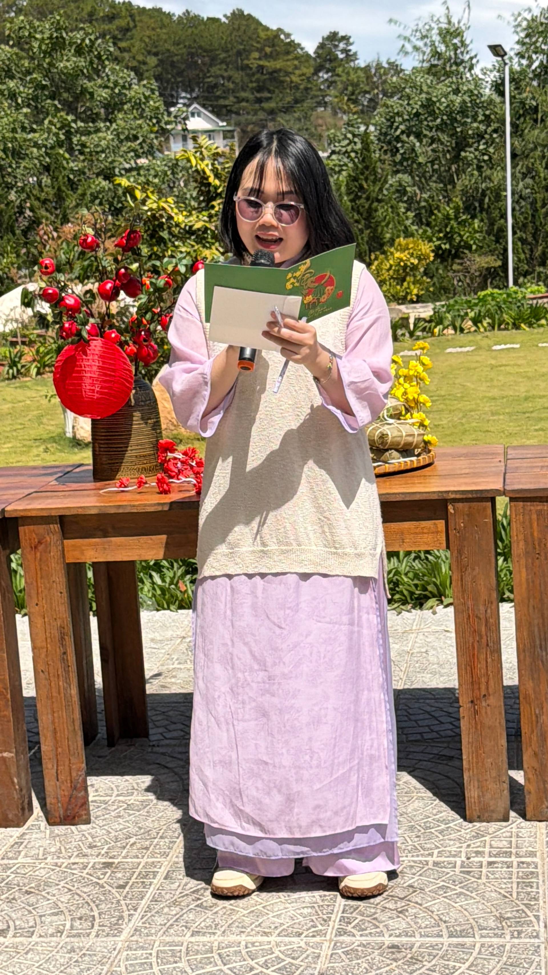 Speaker presenting at Đà Lạt garden event with microphone, reading from program document. Festive outdoor setting decorated with vibrant red roses and Đà Lạt's characteristic pine forest backdrop.