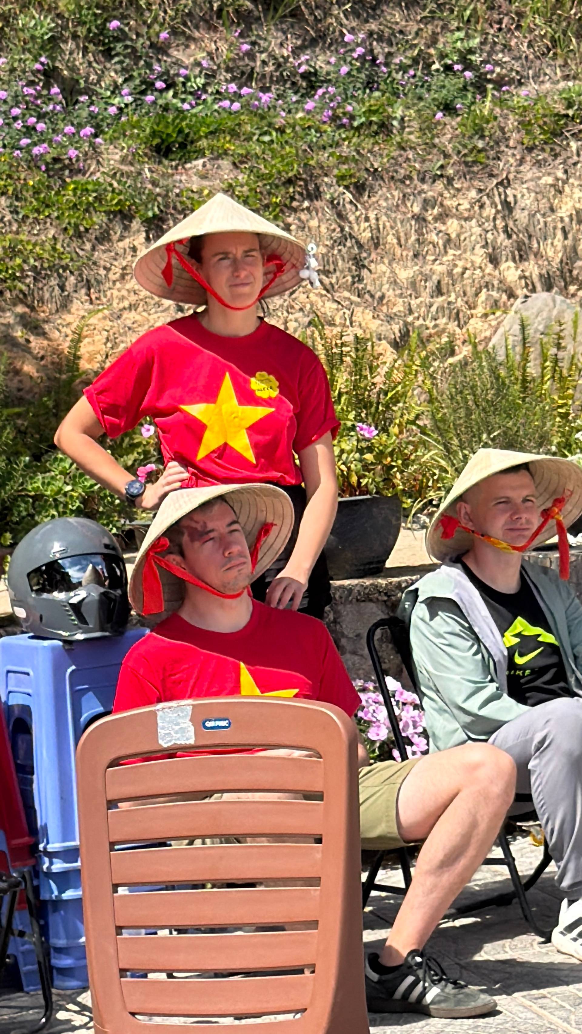 Small group in traditional Vietnamese conical hats gathered for cultural celebration in Đà Lạt garden. Festive outdoor scene with vibrant purple flowers and patriotic red-and-yellow themed clothing.