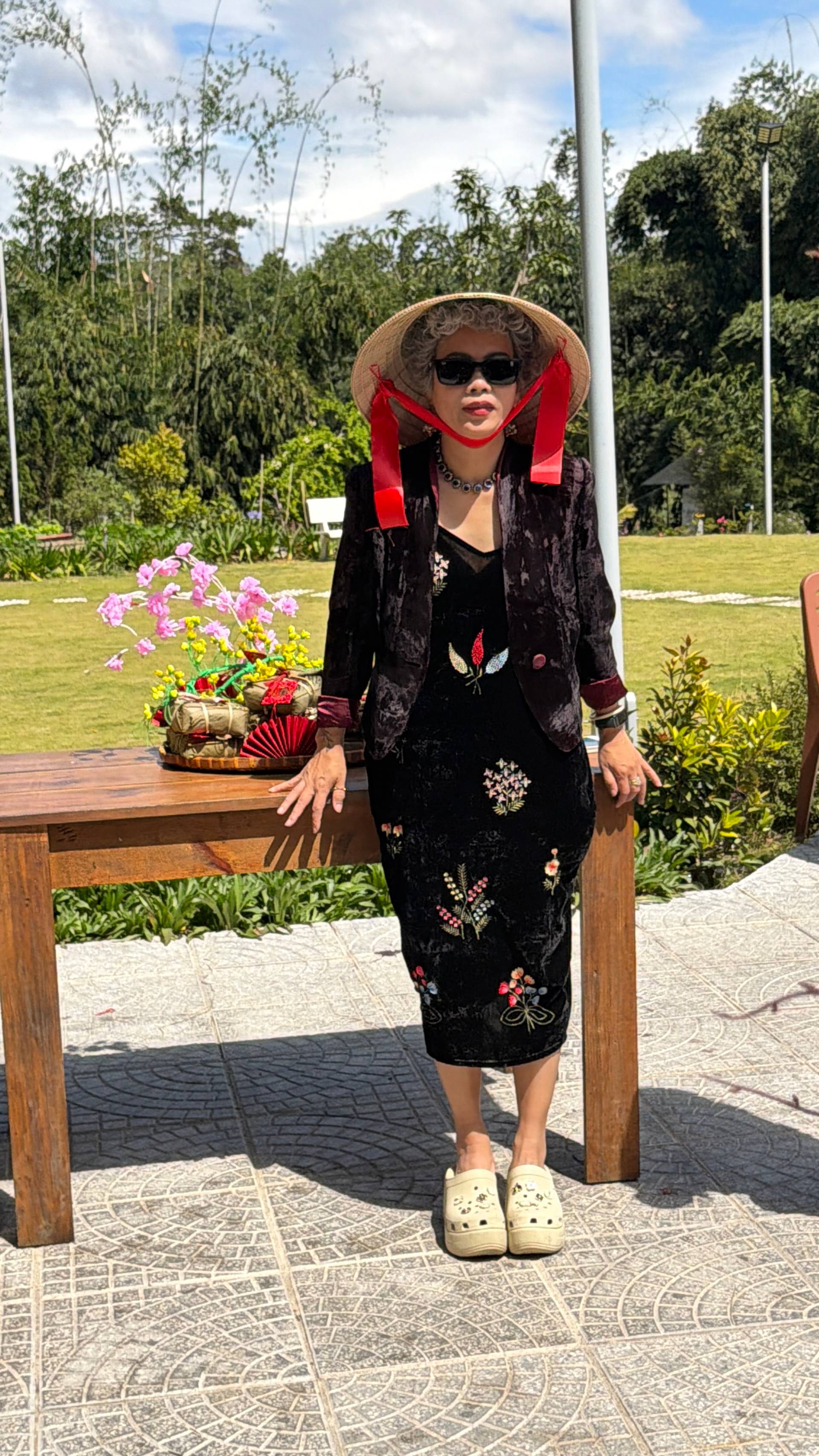 Solo portrait at traditional Vietnamese garden event in Đà Lạt featuring conical hat and ornate traditional costume. Participant stands by decorative floral table with pink flowers against backdrop of misty pine forest.