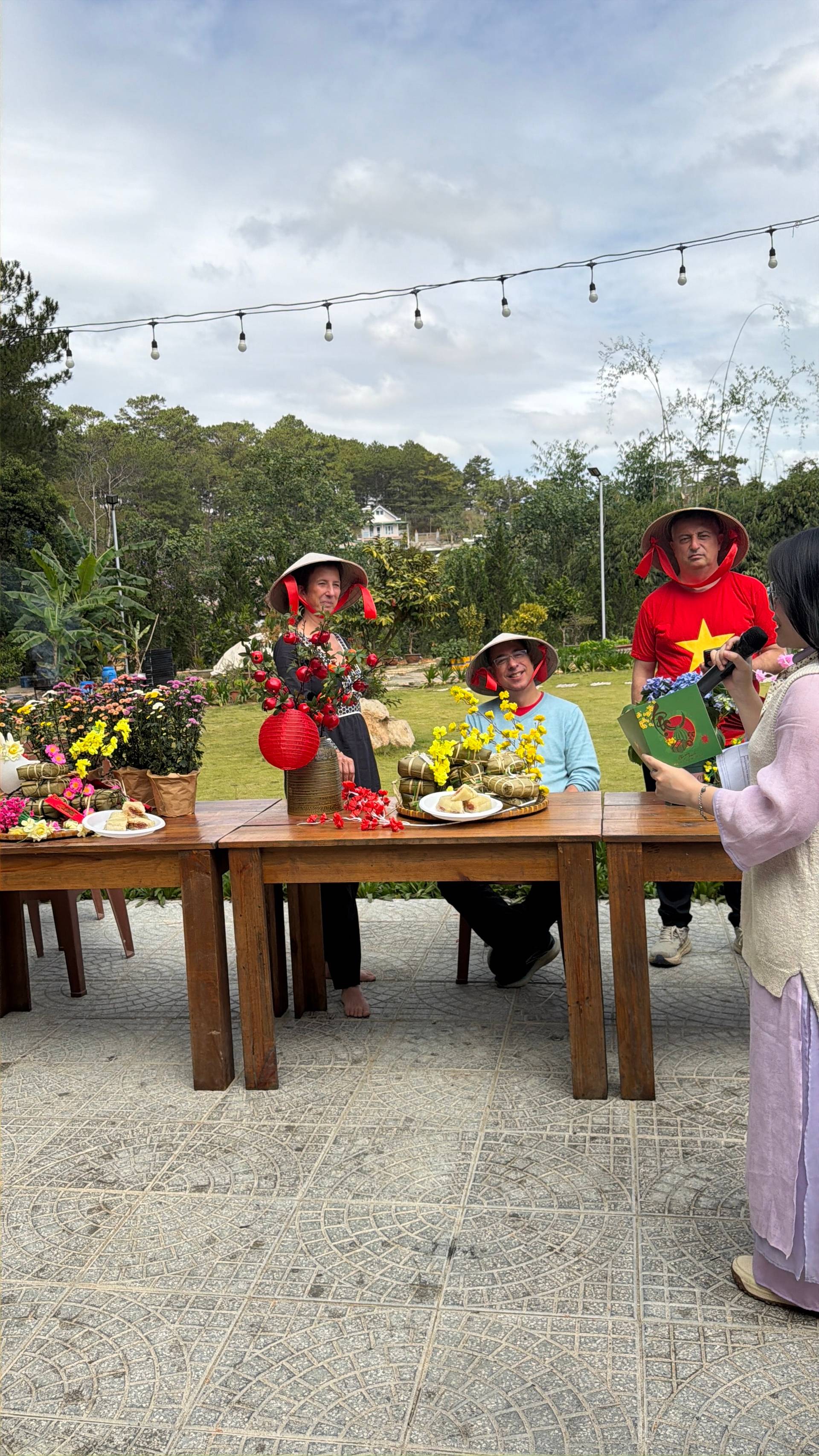Vibrant cultural celebration in Đà Lạt featuring guests in traditional Vietnamese áo dài and conical hats. Festive garden event decorated with red and yellow flowers, paper lanterns, and string lights overlooking pine-forested landscape.