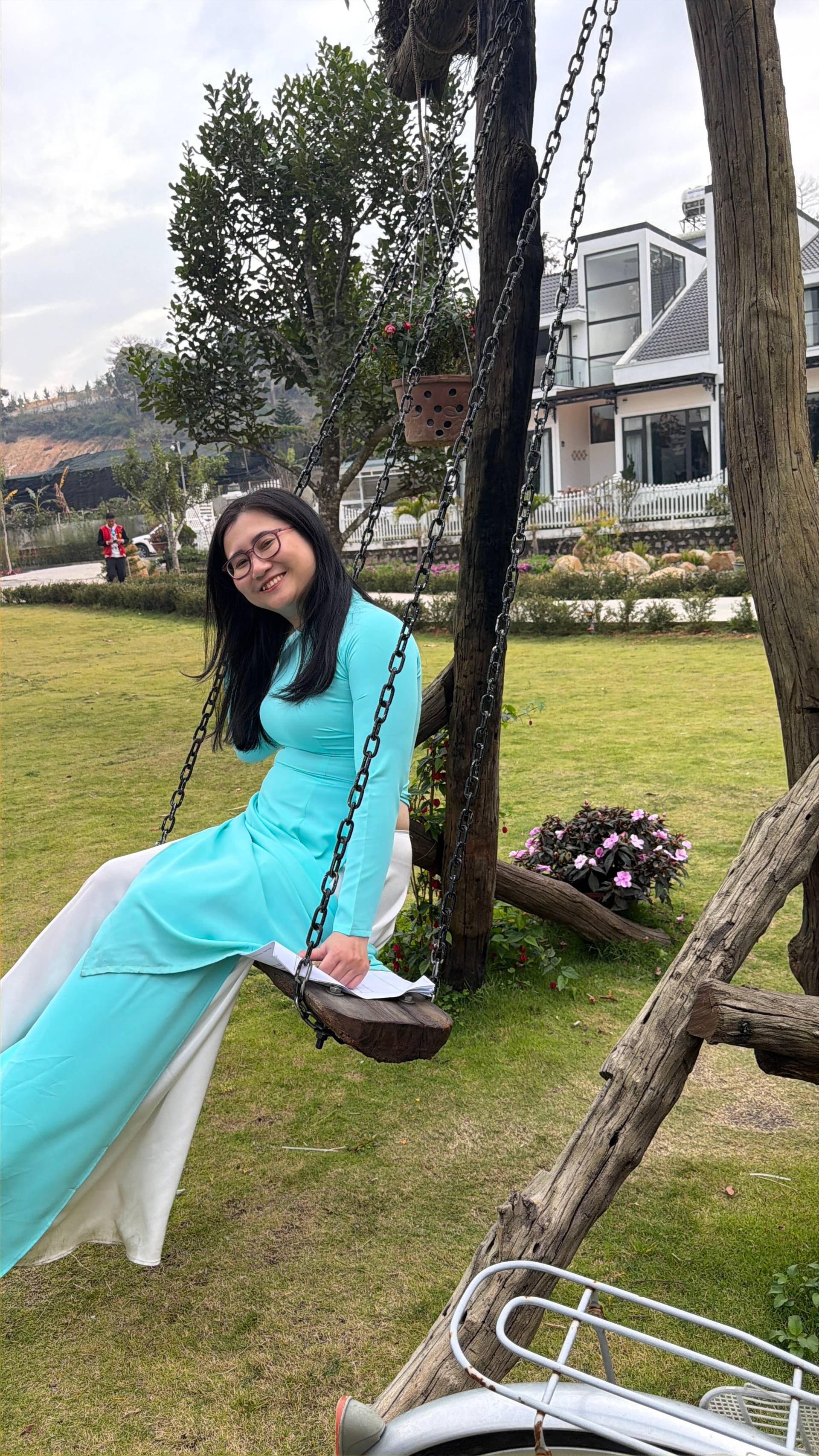 Person in traditional Vietnamese áo dài on a rustic tree swing at a Đà Lạt villa garden, surrounded by pink flowers and modern architecture in a peaceful countryside setting.