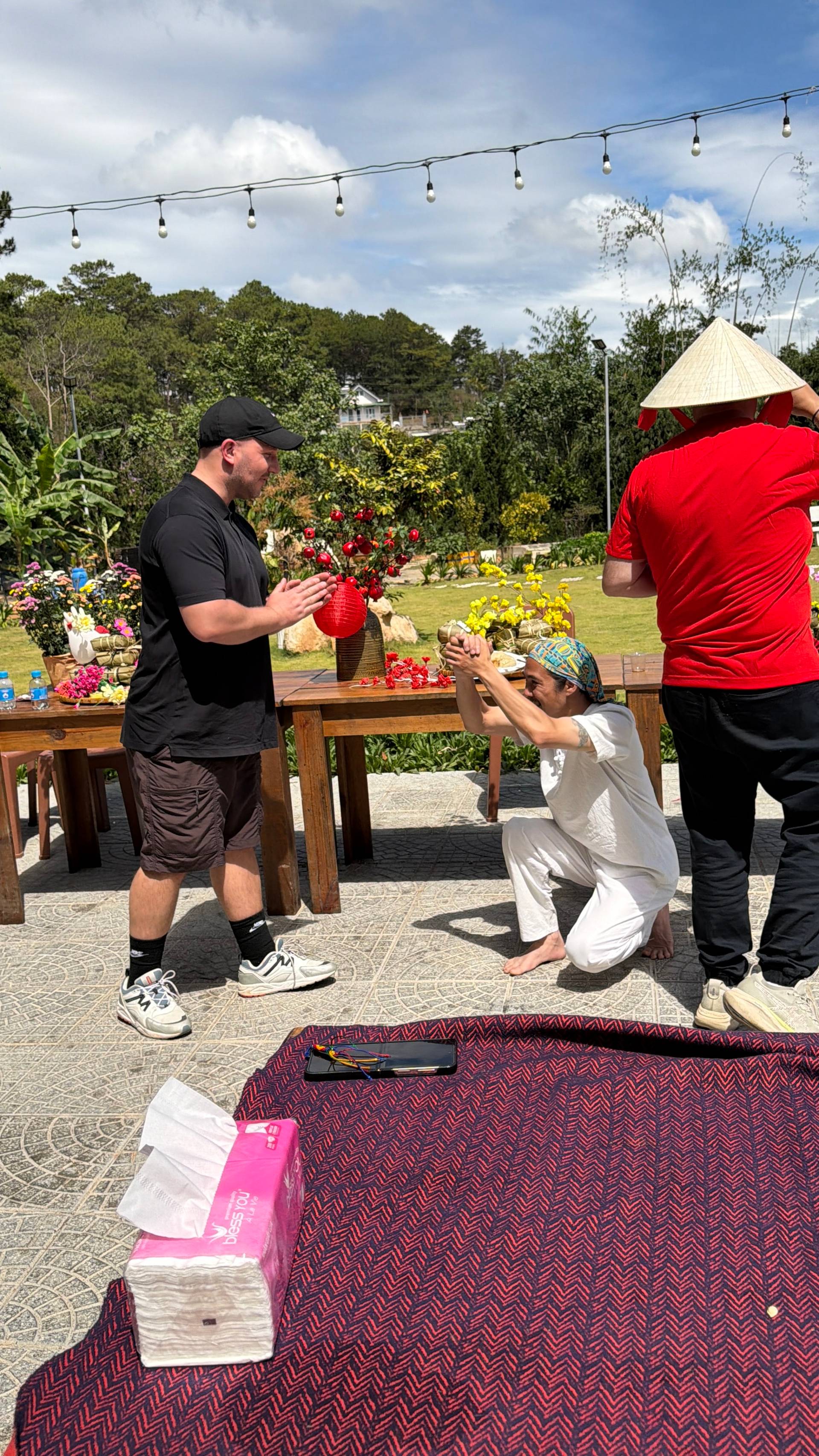 Interactive lantern-making activity at a Đà Lạt garden event with colorful flowers and traditional conical hats in a bright, sunny hillside setting.