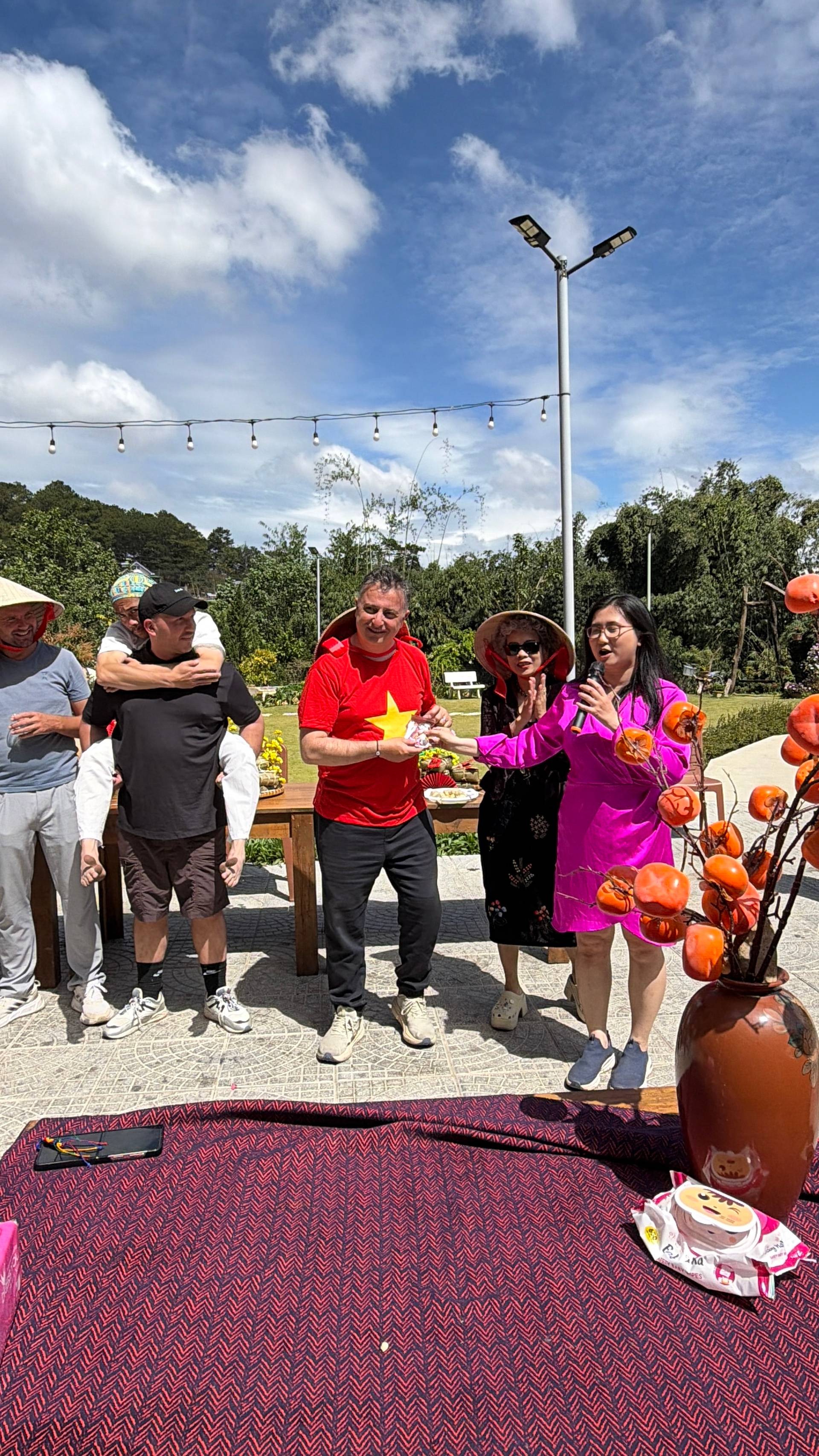 A playful and joyful moment at a Đà Lạt cultural event featuring someone proudly displaying the massive traditional Vietnamese conical hat (nón lá). The outdoor garden setting with fresh local foods captures the spirit of Vietnamese hospitality and celebration.