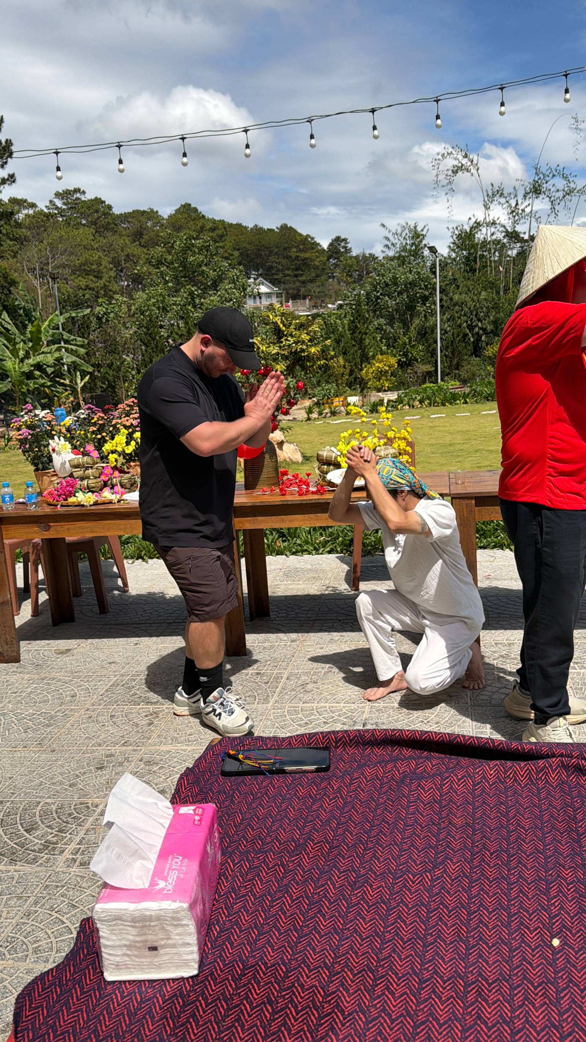 A vibrant cultural heritage celebration in Đà Lạt's lush mountain setting, featuring a diverse group of participants in traditional dress. The festive daytime gathering showcases red and gold star motifs alongside natural pine forest vistas.