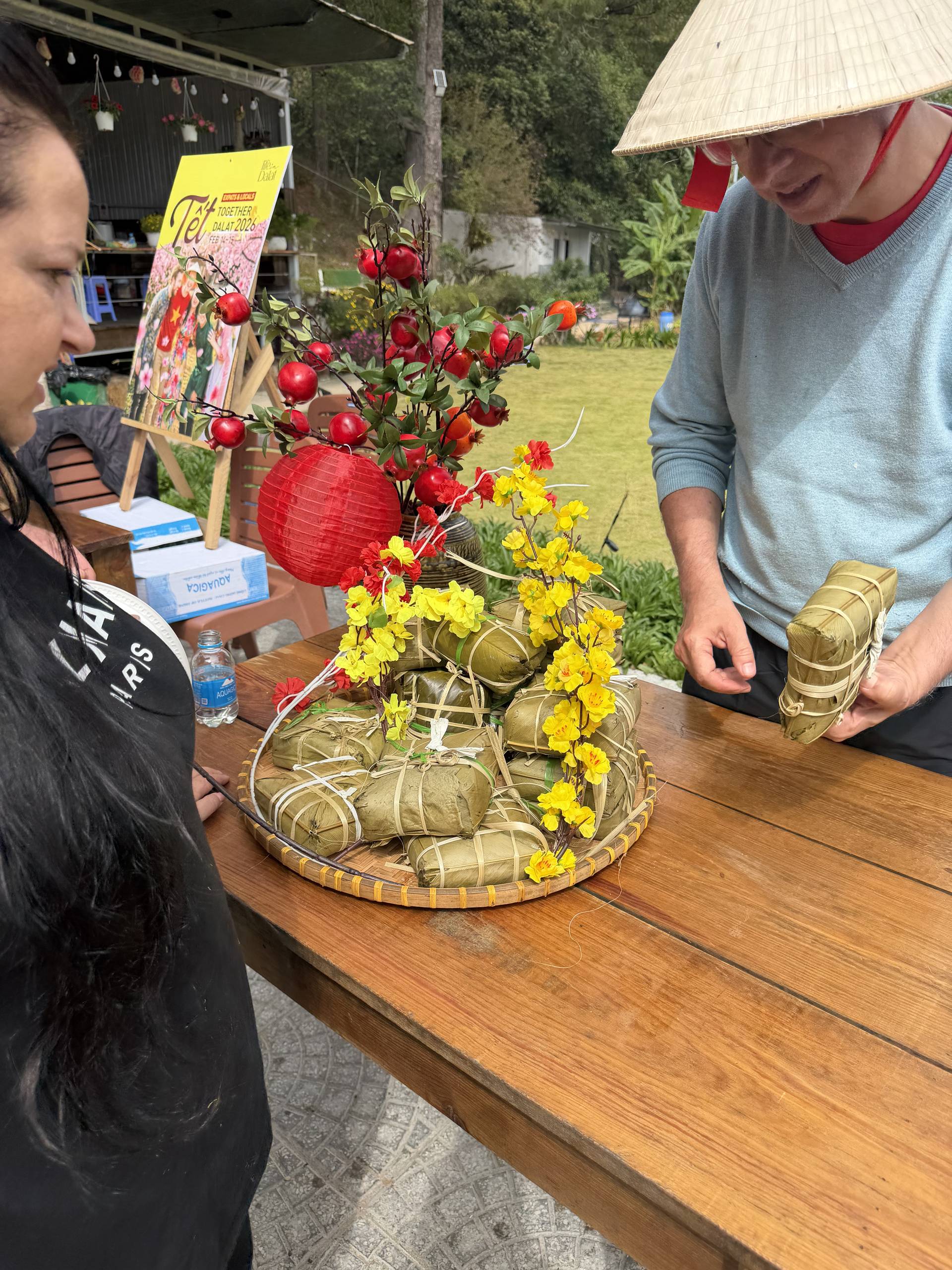 A vibrant daytime cultural event in Đà Lạt's pine forest setting showcasing traditional Vietnamese craftsmanship and heritage. The display features abundant red lanterns, colorful dahlias, and traditional woven elements at an outdoor wooden table venue.