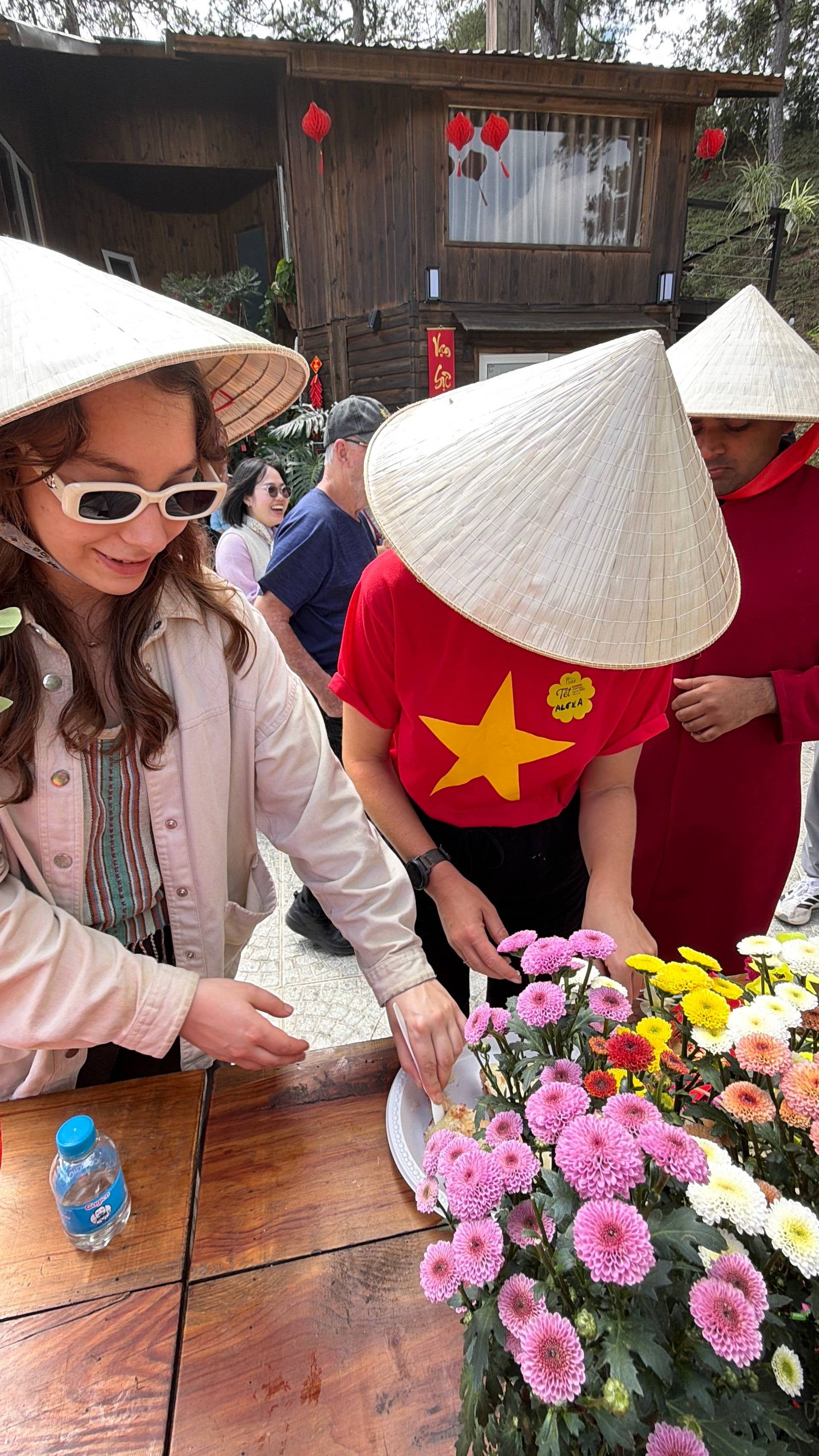 A festive outdoor daytime celebration in Đà Lạt featuring participants in traditional Vietnamese attire with red and gold star symbols. The mountain garden venue displays vibrant dahlias, decorative lanterns, and showcases Vietnamese cultural pride in a pine-forested setting.