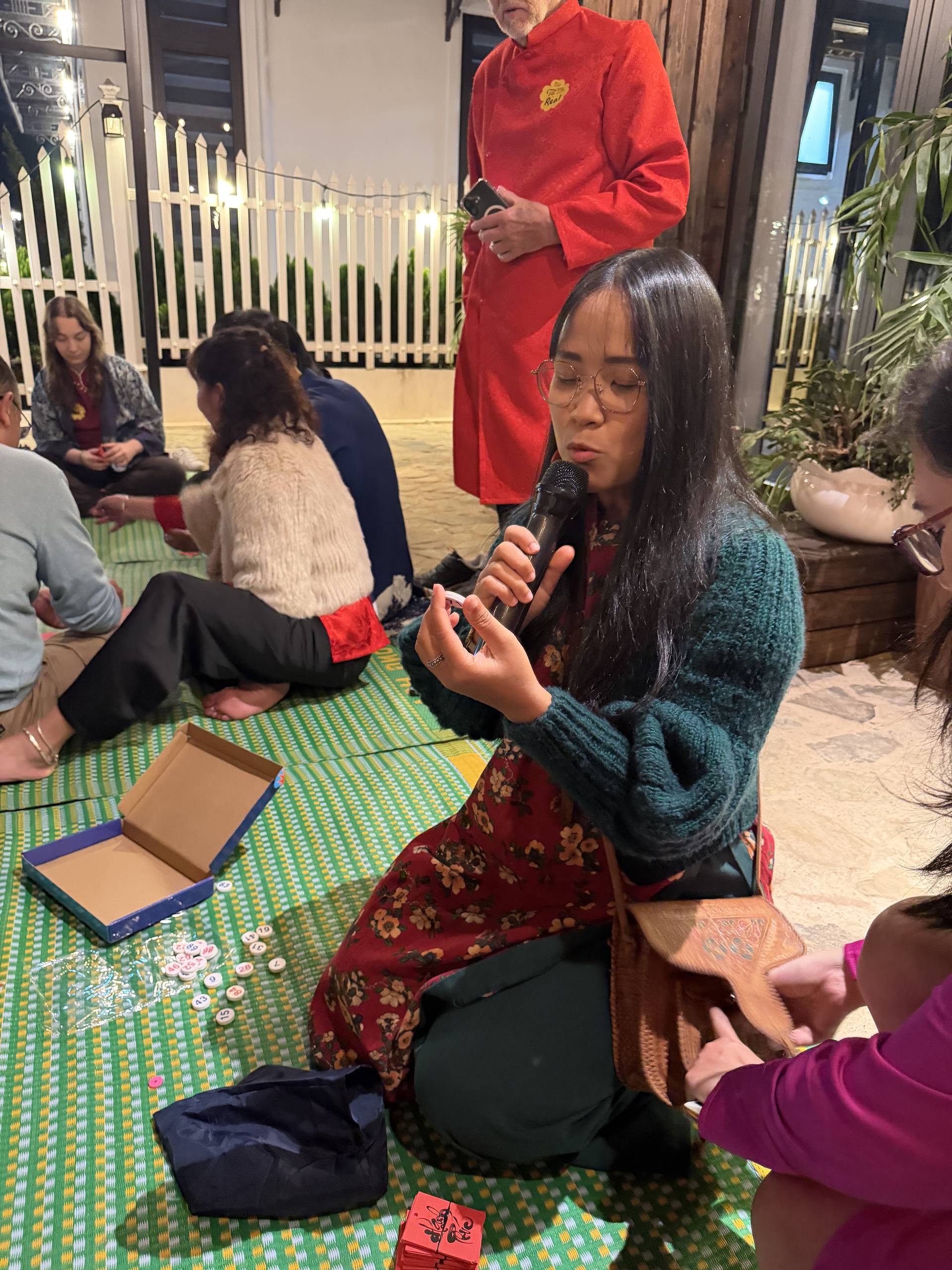 An indoor cultural event in Đà Lạt with someone presenting over a colorful traditional fabric mat, wearing red ceremonial clothing, surrounded by audience members.