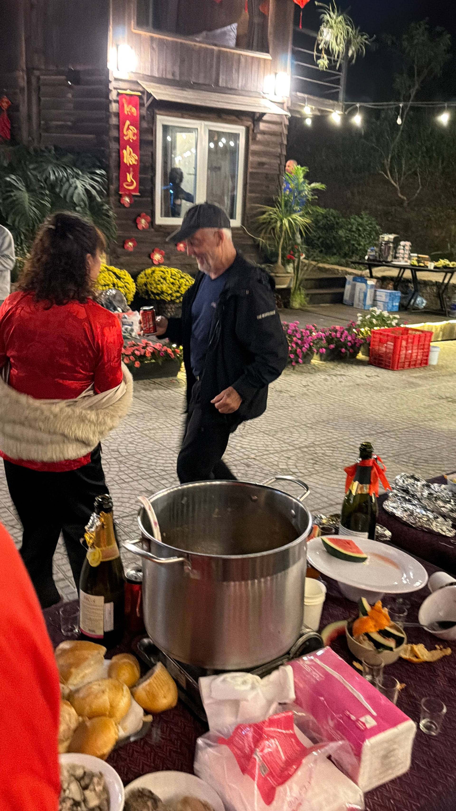 A festive nighttime Lunar New Year celebration at a Đà Lạt venue with a large communal cooking pot, traditional red lanterns, and golden chrysanthemums creating a warm holiday atmosphere.