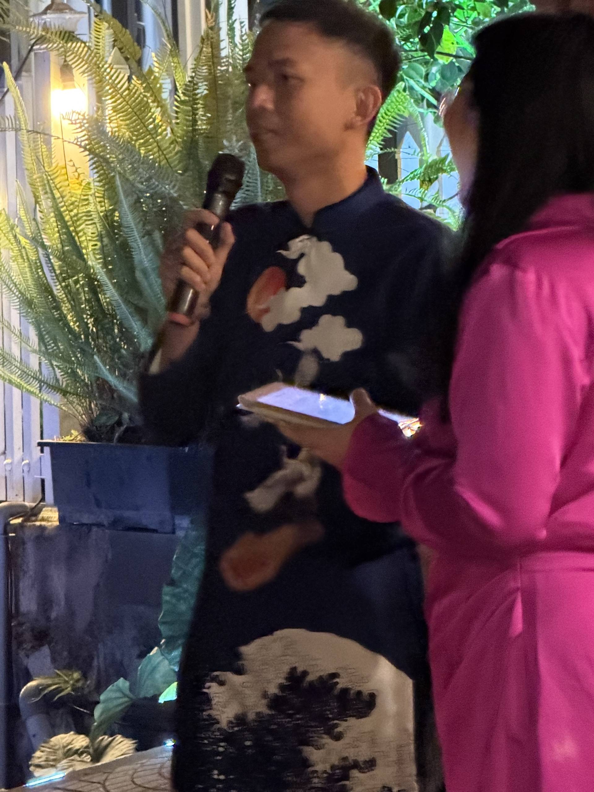 An indoor presentation or performance at a Đà Lạt event surrounded by lush hanging ferns and tropical plants, with an attendee taking notes during the presentation.