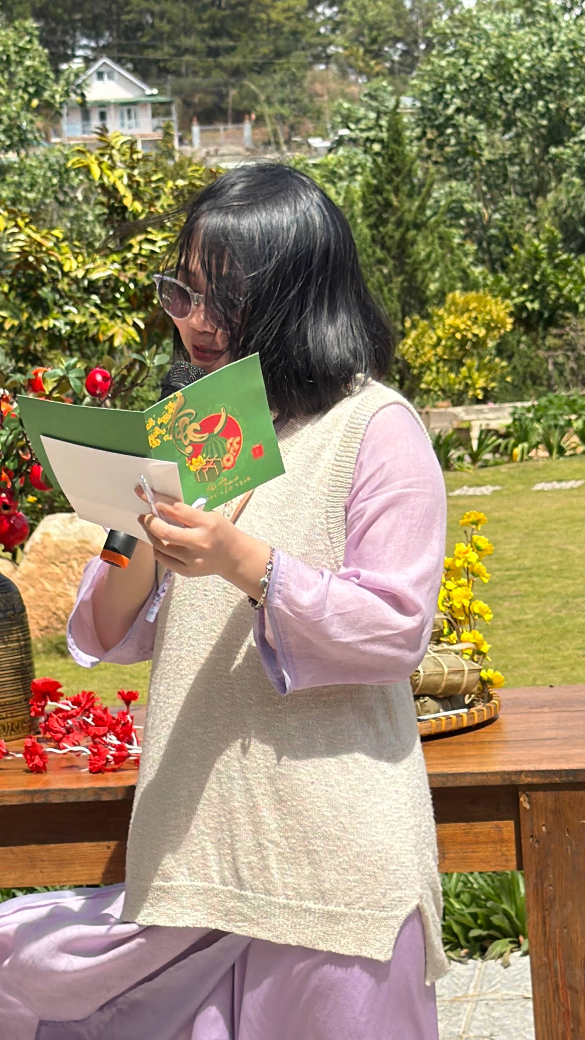 A serene garden reading moment in Đà Lạt featuring lush flowers and pine-forested hills, where someone quietly enjoys a document or invitation at a scenic outdoor venue.