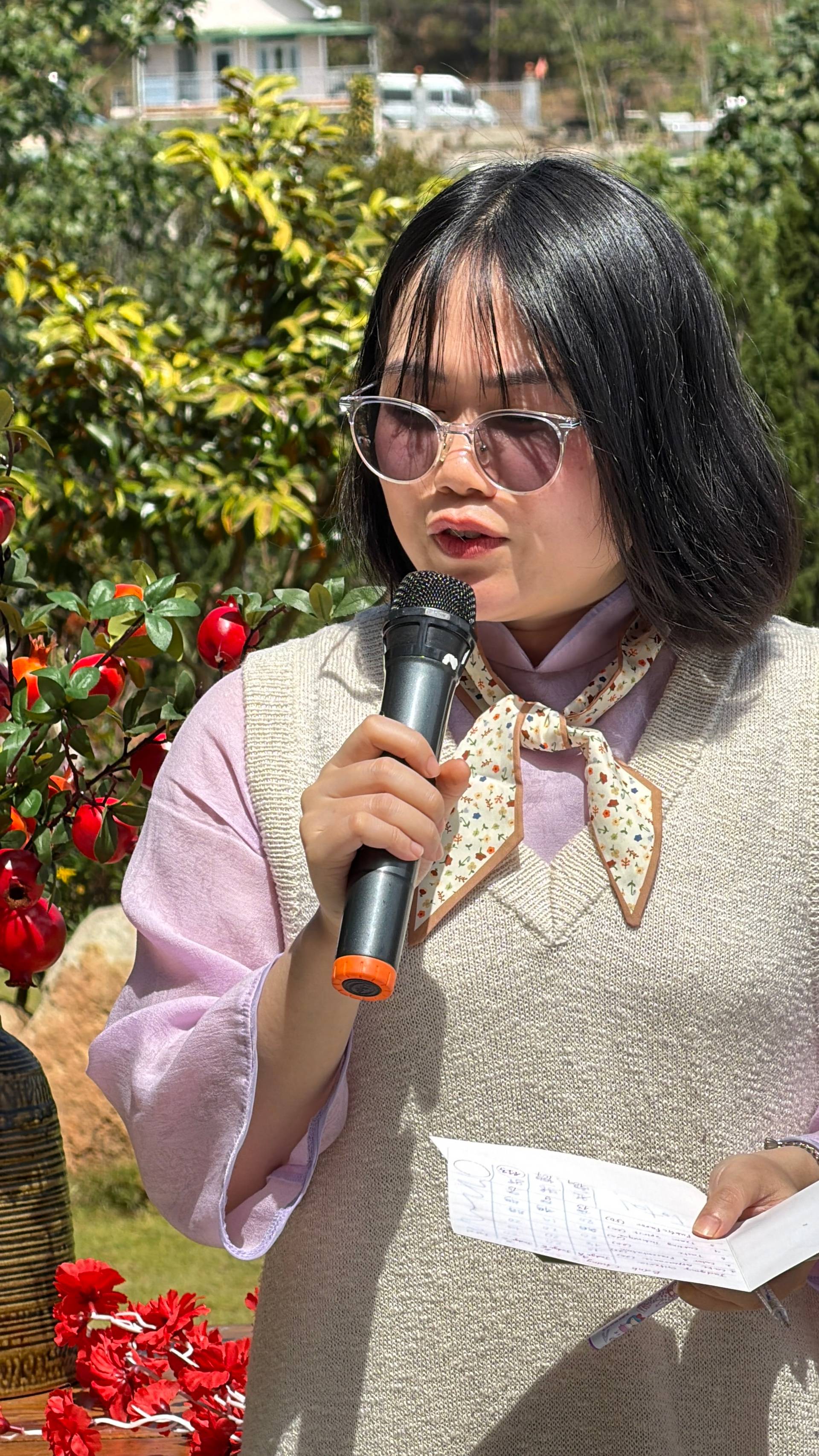 Outdoor garden speaker at a Đà Lạt cultural event, wearing traditional conical hat, with vibrant red flowers and event program visible.