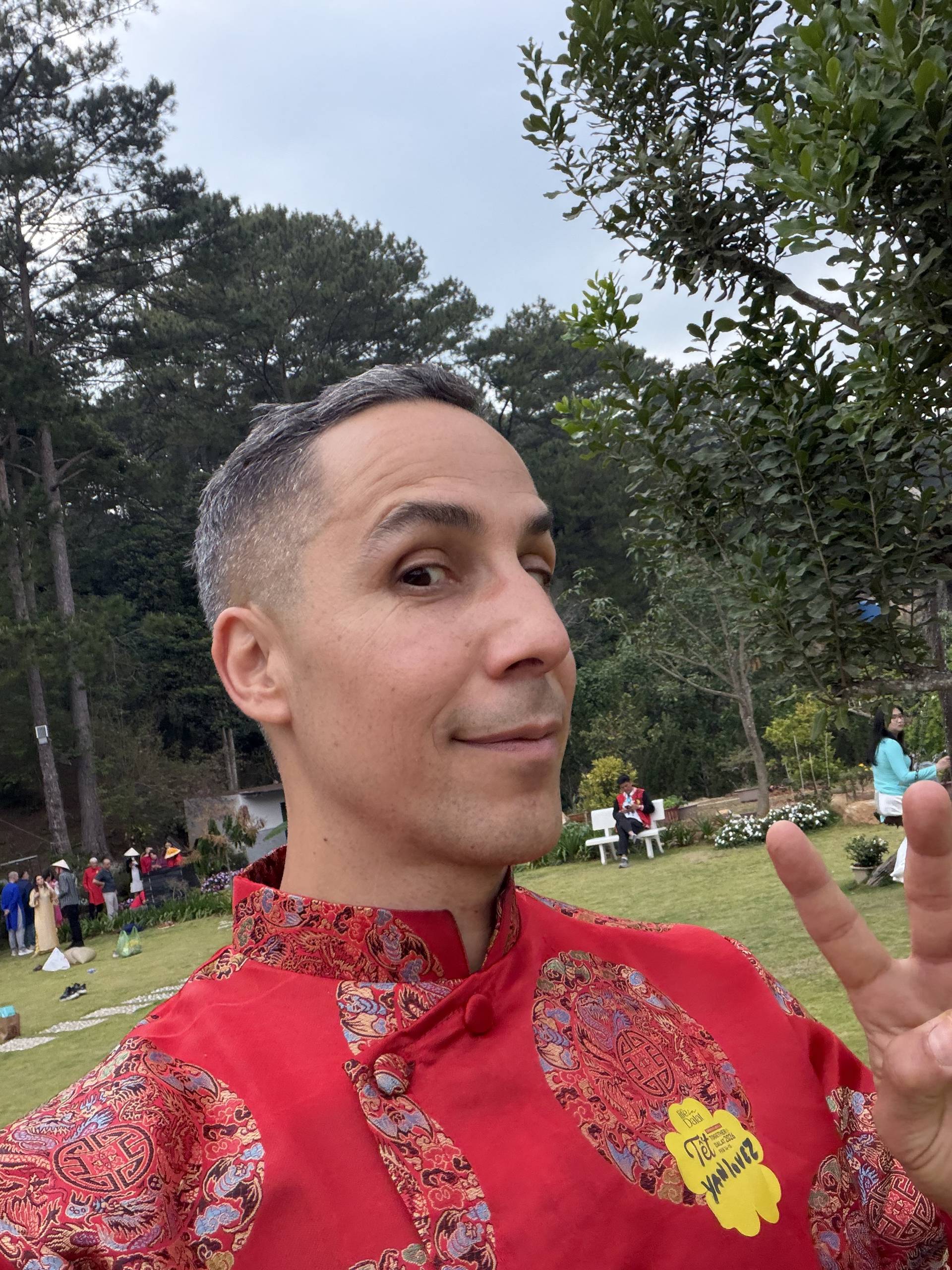 Outdoor speaker at a Đà Lạt event, wearing traditional conical hat and red formal attire, with pine forest and grassy venue backdrop.
