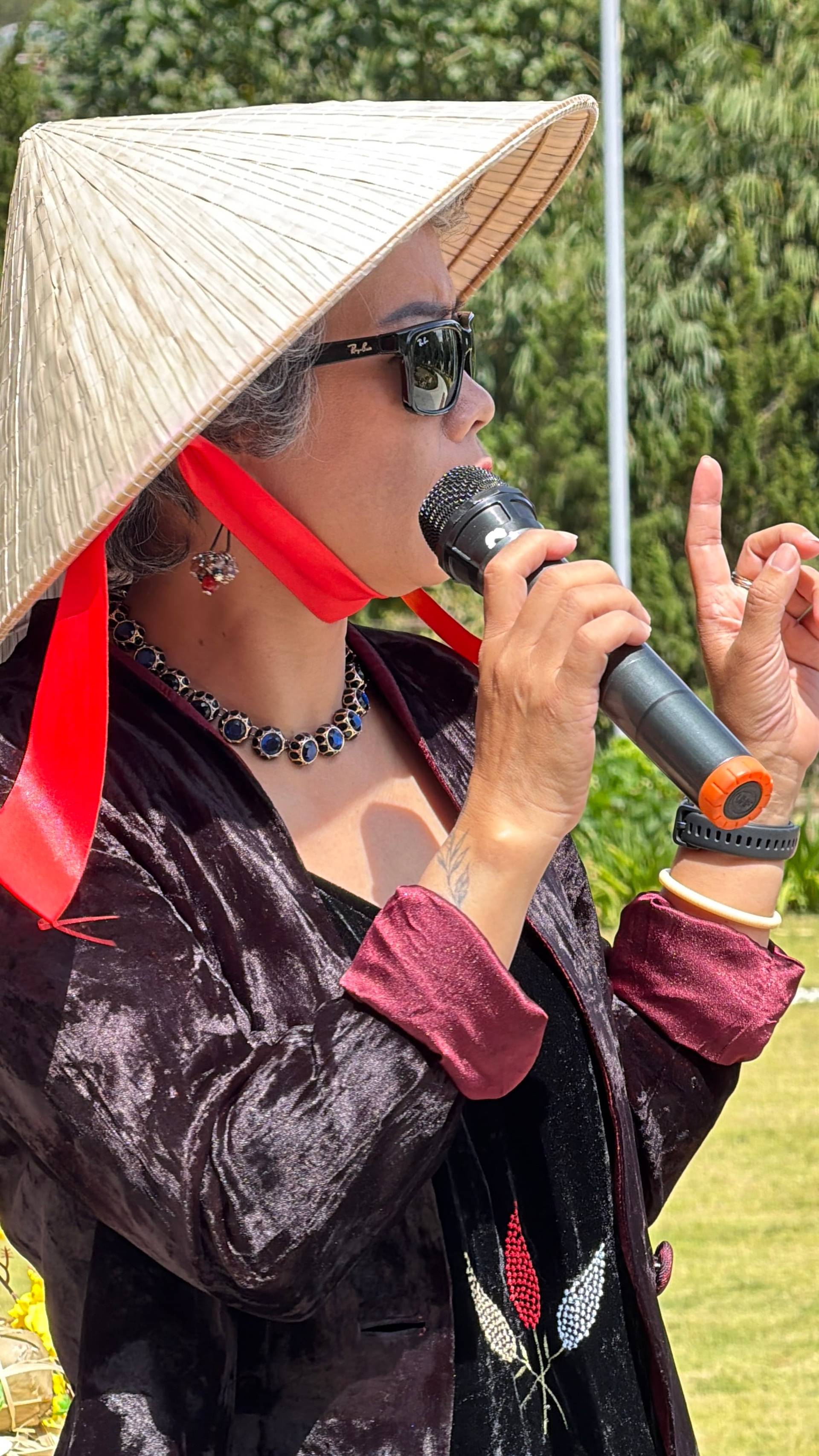 Daytime performer speaking at a microphone in Đà Lạt's green forest setting, wearing traditional red formal attire and conical hat.