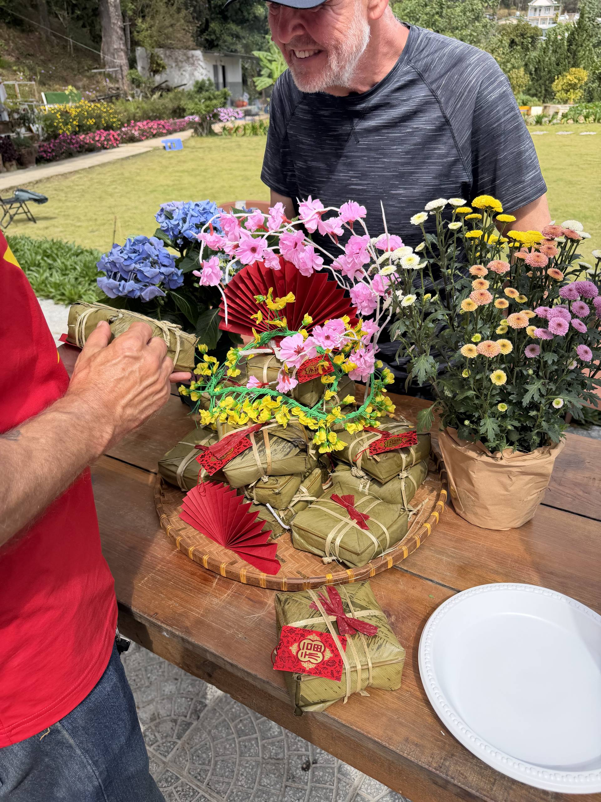 Garden celebration in Đà Lạt featuring vibrant floral displays with traditional red Vietnamese fan decorations, wrapped gifts with red ribbons, and colorful bouquets of hydrangeas, daisies, and chrysanthemums.
