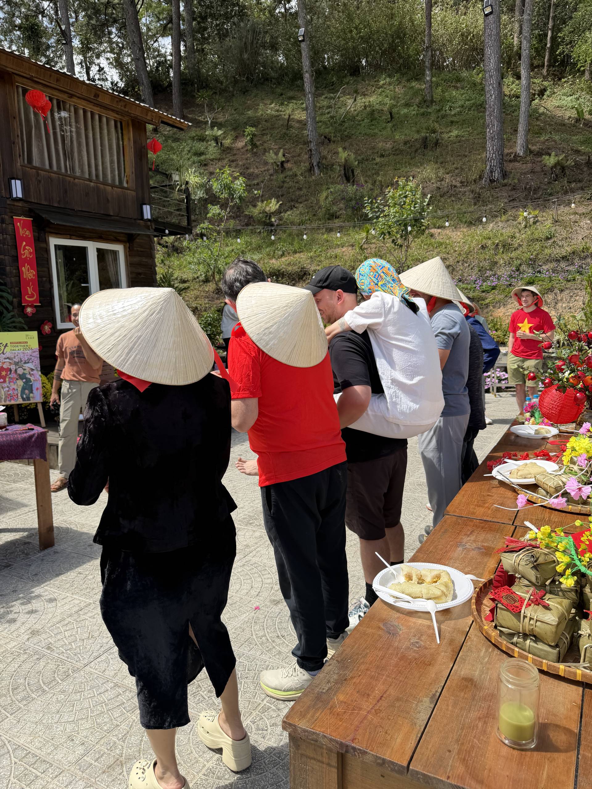 Community gathering at a wooden pavilion in Đà Lạt's forested setting with visitors in conical hats enjoying refreshments and food around decorated tables.