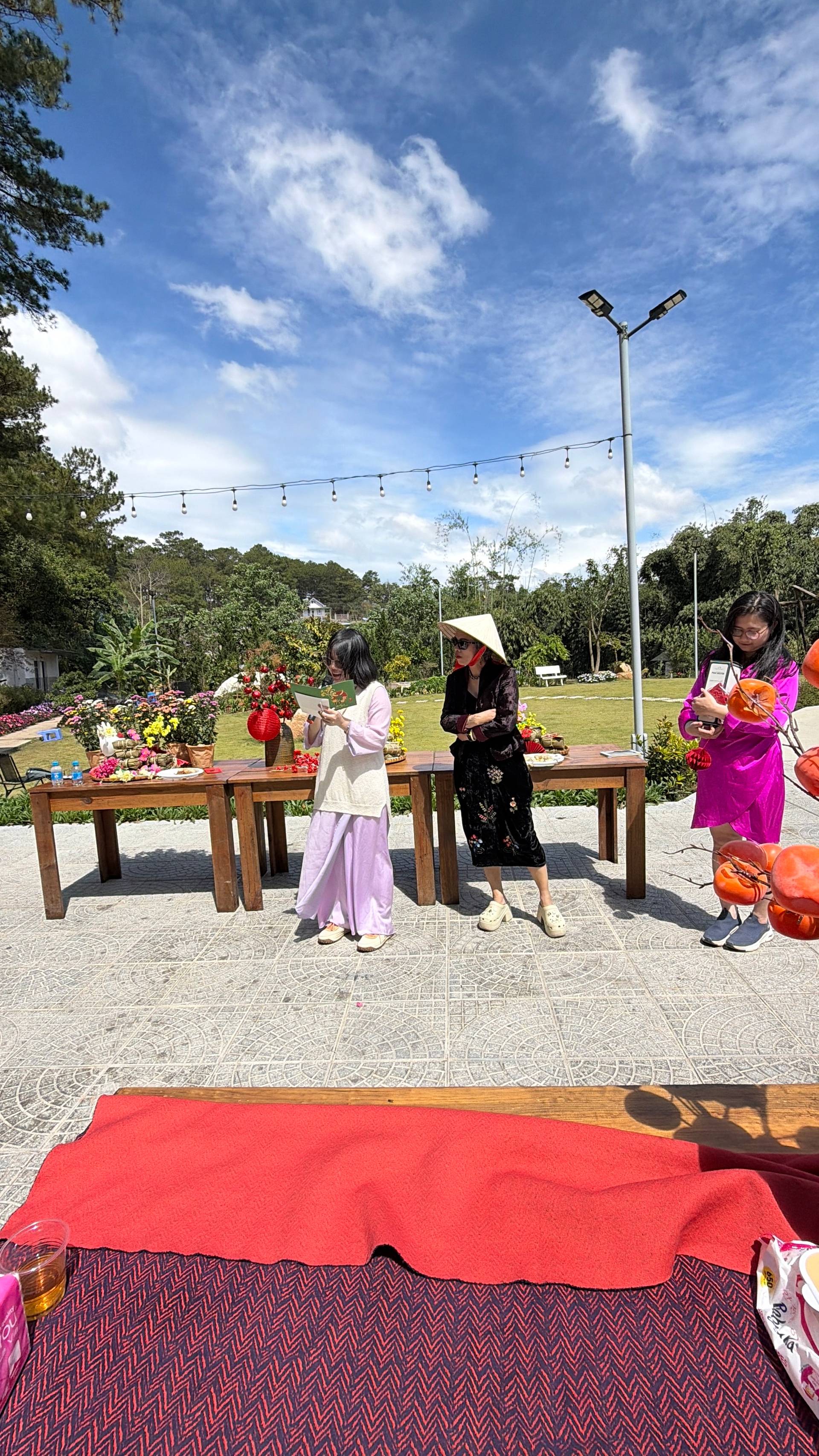 Large outdoor community celebration in Đà Lạt countryside with visitors gathered at wooden tables surrounded by vibrant flowers, string lights, and forested hillsides under clear blue sky.