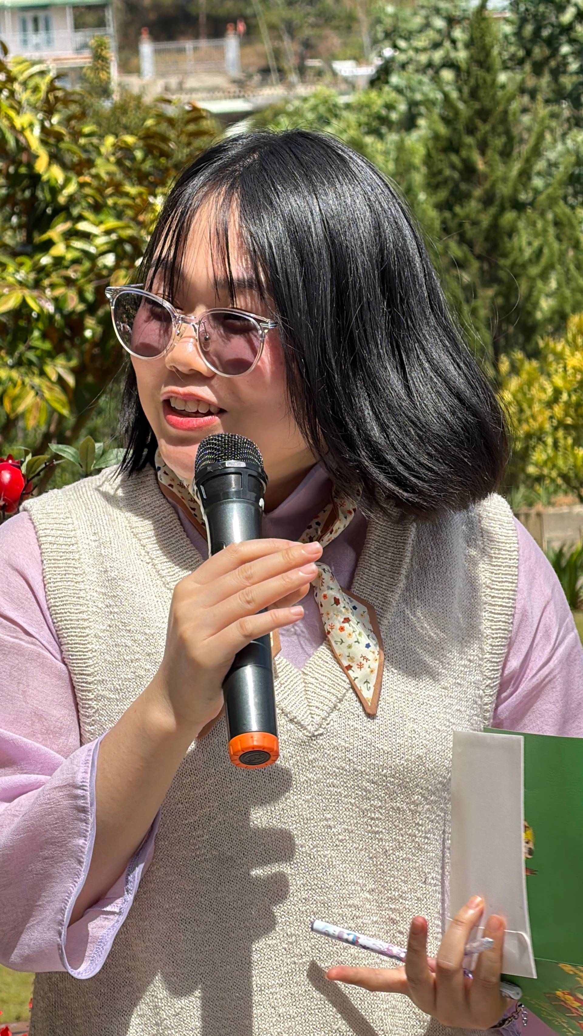 Daytime presentation or speech at an outdoor event venue in Đà Lạt with coniferous forest setting visible behind. Speaker addressing attendees in an open garden space with landscaping and traditional buildings in background.
